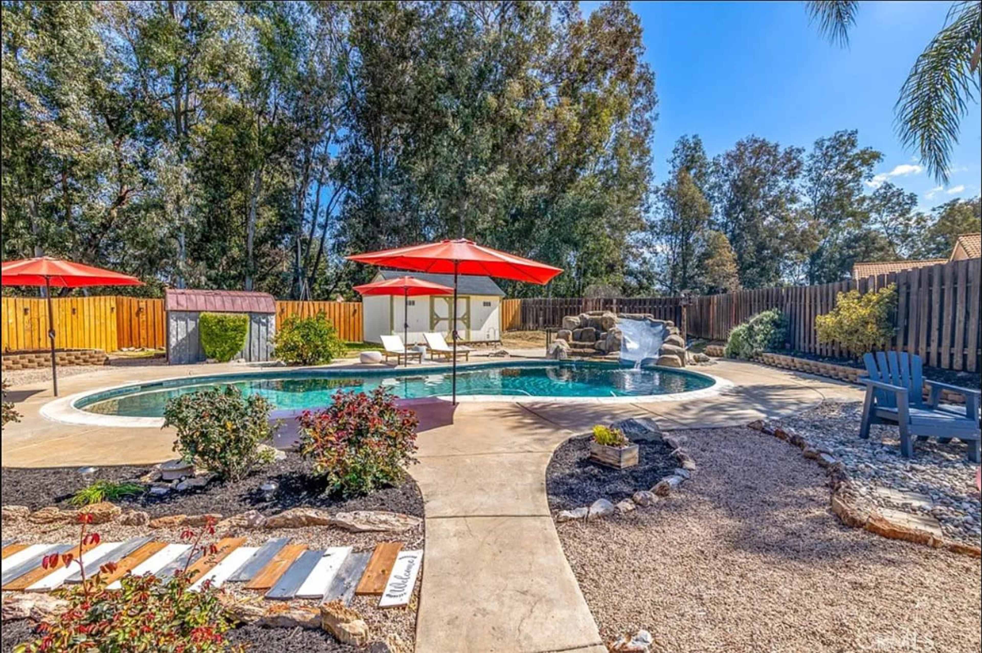 A backyard pool area surrounded by landscaped gardens, featuring two red umbrellas over lounge chairs and a rock waterfall.