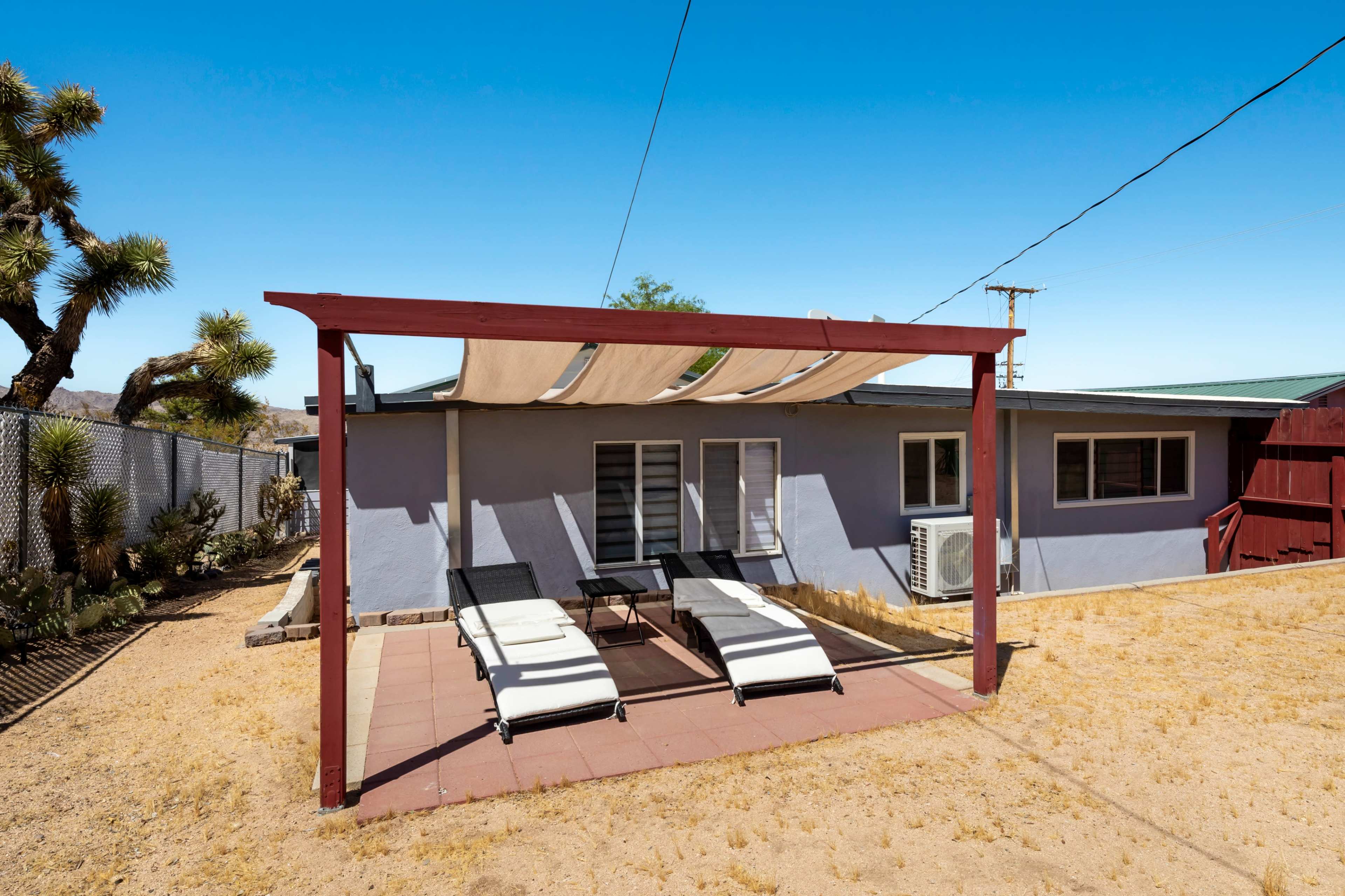 A shaded patio area with two lounge chairs sits beside a grey house in a dry, desert landscape.
