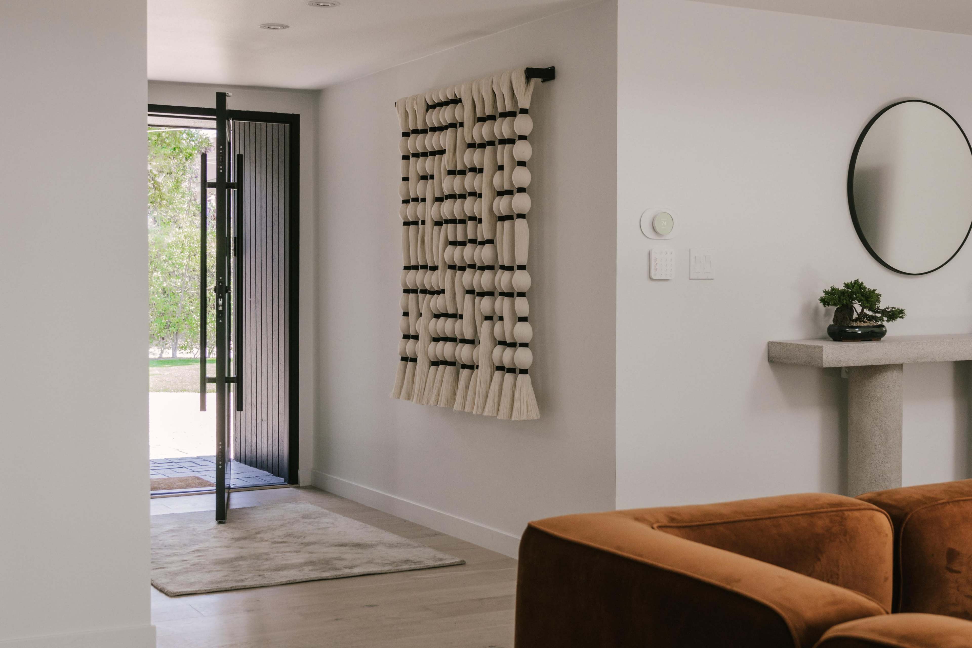 A modern entryway features an open door leading outside, a textile wall hanging, a round mirror, and a small plant on a console table.