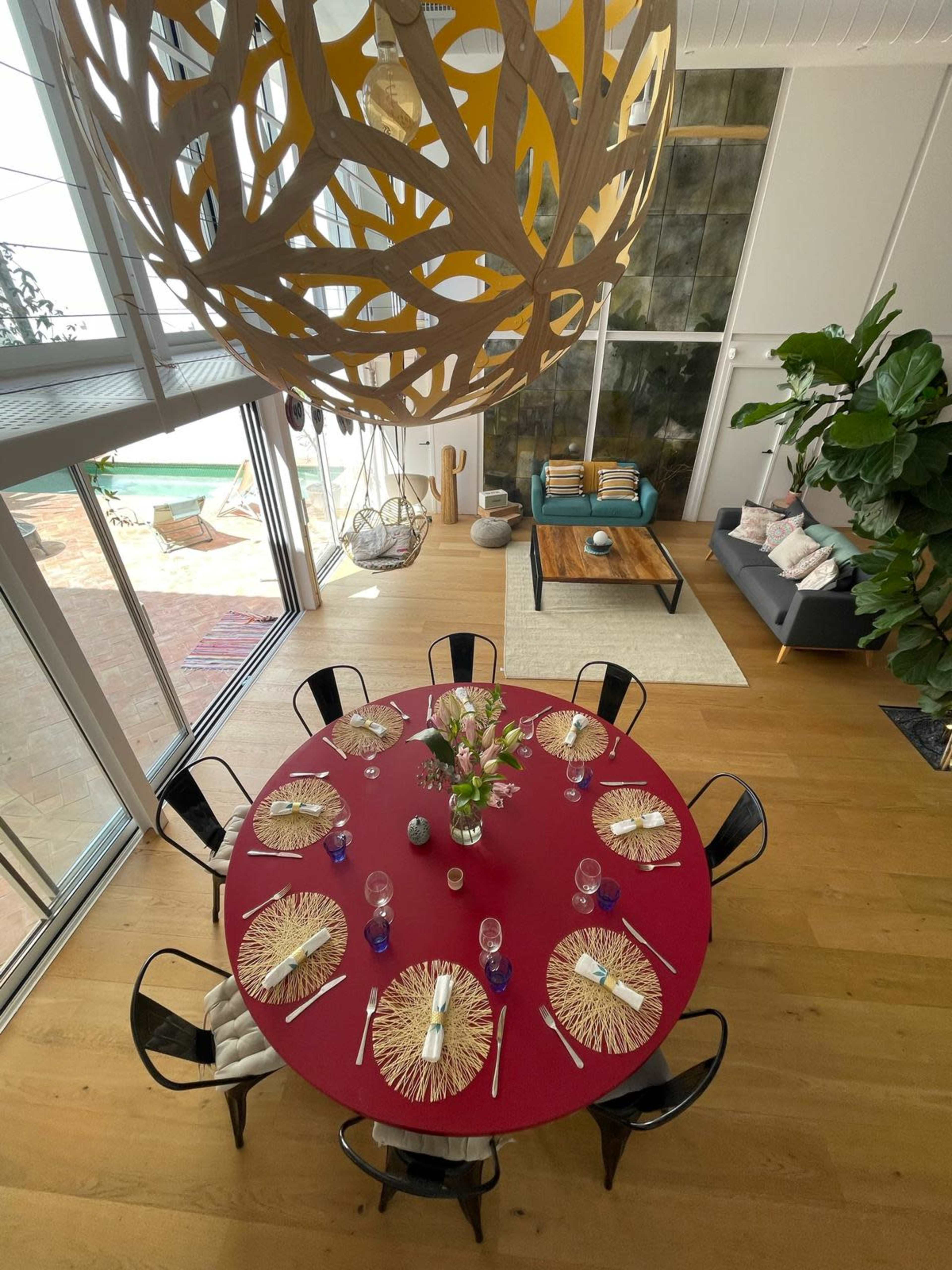 A large round dining table is set with decorative placemats and flower centerpieces, surrounded by black chairs, with a bright chandelier overhead and a view of a pool and living area in the background.