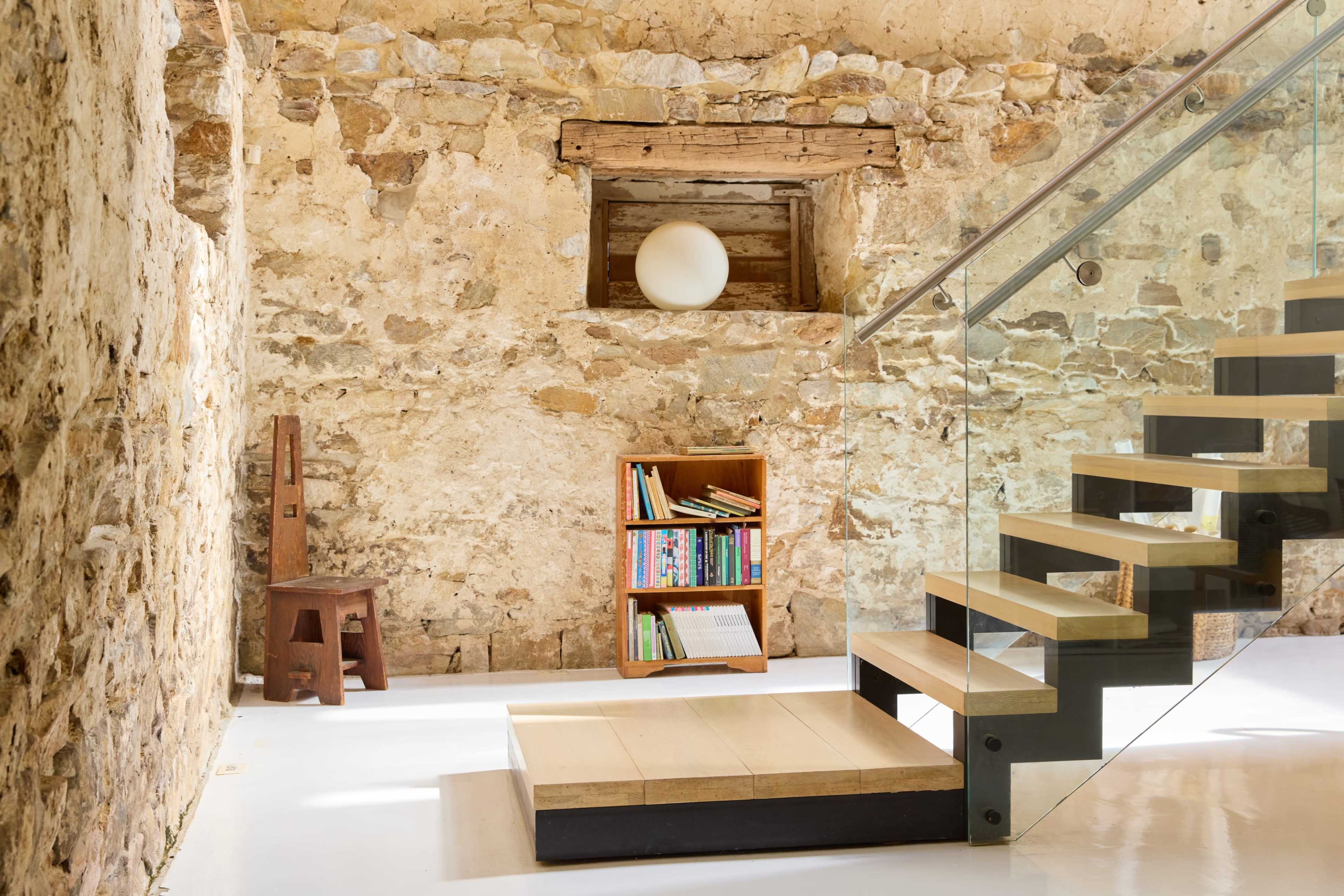The image shows a modern interior with a glass staircase leading up to a small window set into a stone wall, beside a wooden bookshelf filled with books.