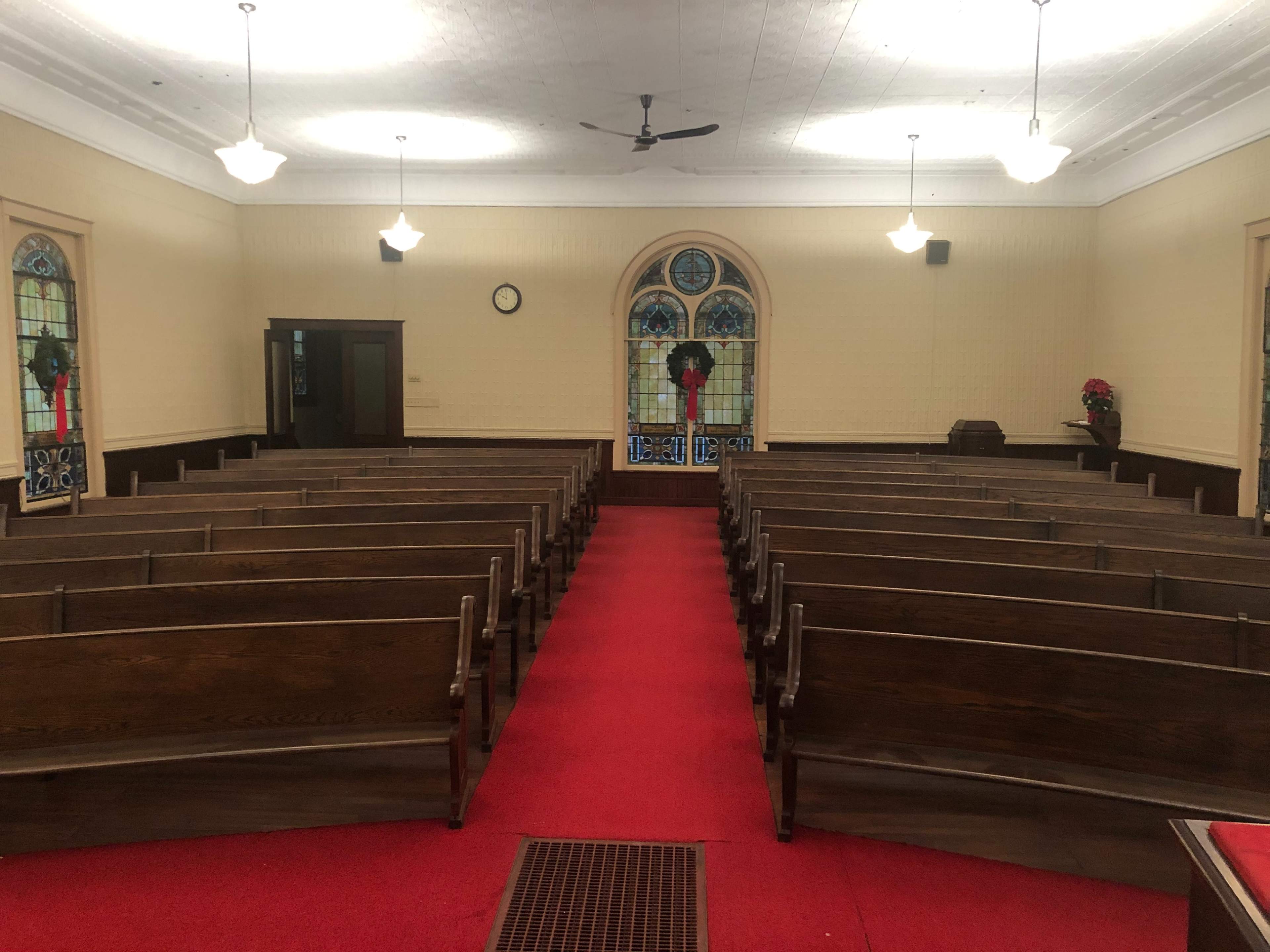 The interior of a church with wooden pews arranged in rows along a red carpet aisle, and stained glass windows at the front.
