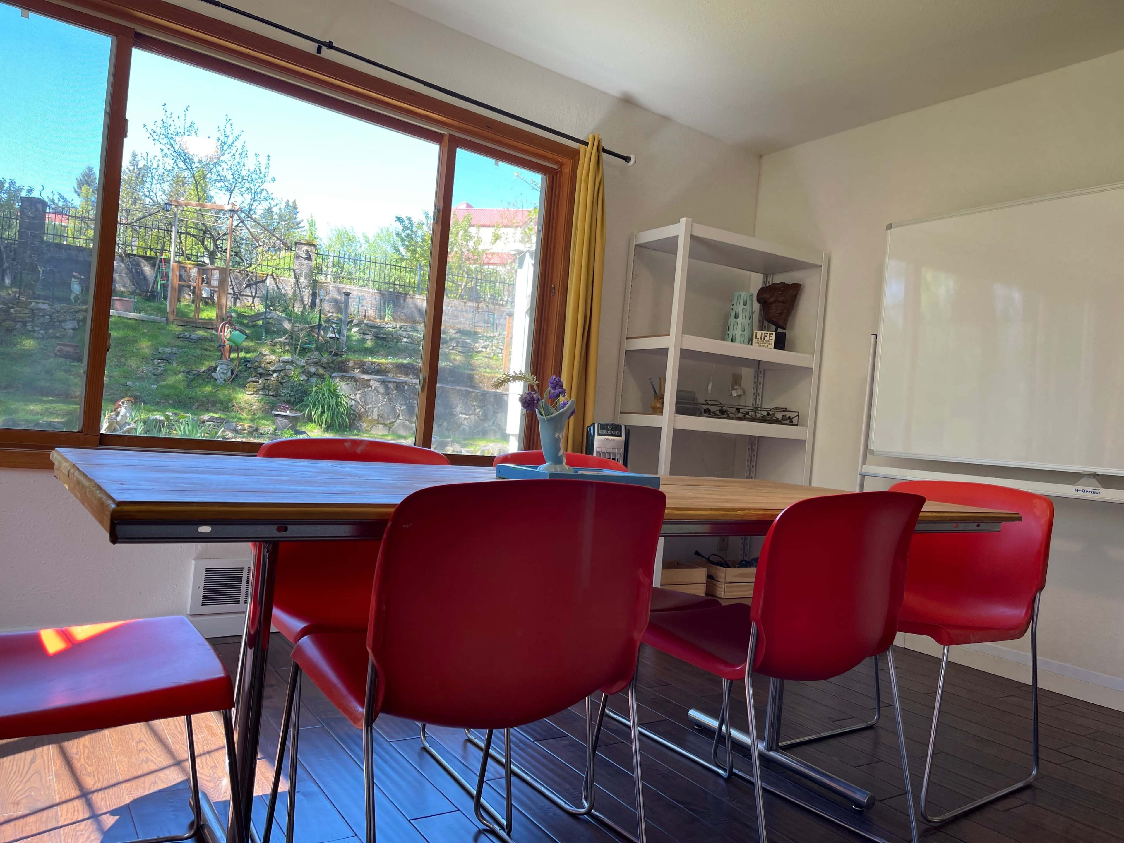 A modern meeting room features a wooden table surrounded by red chairs, with a large window overlooking a landscaped garden.