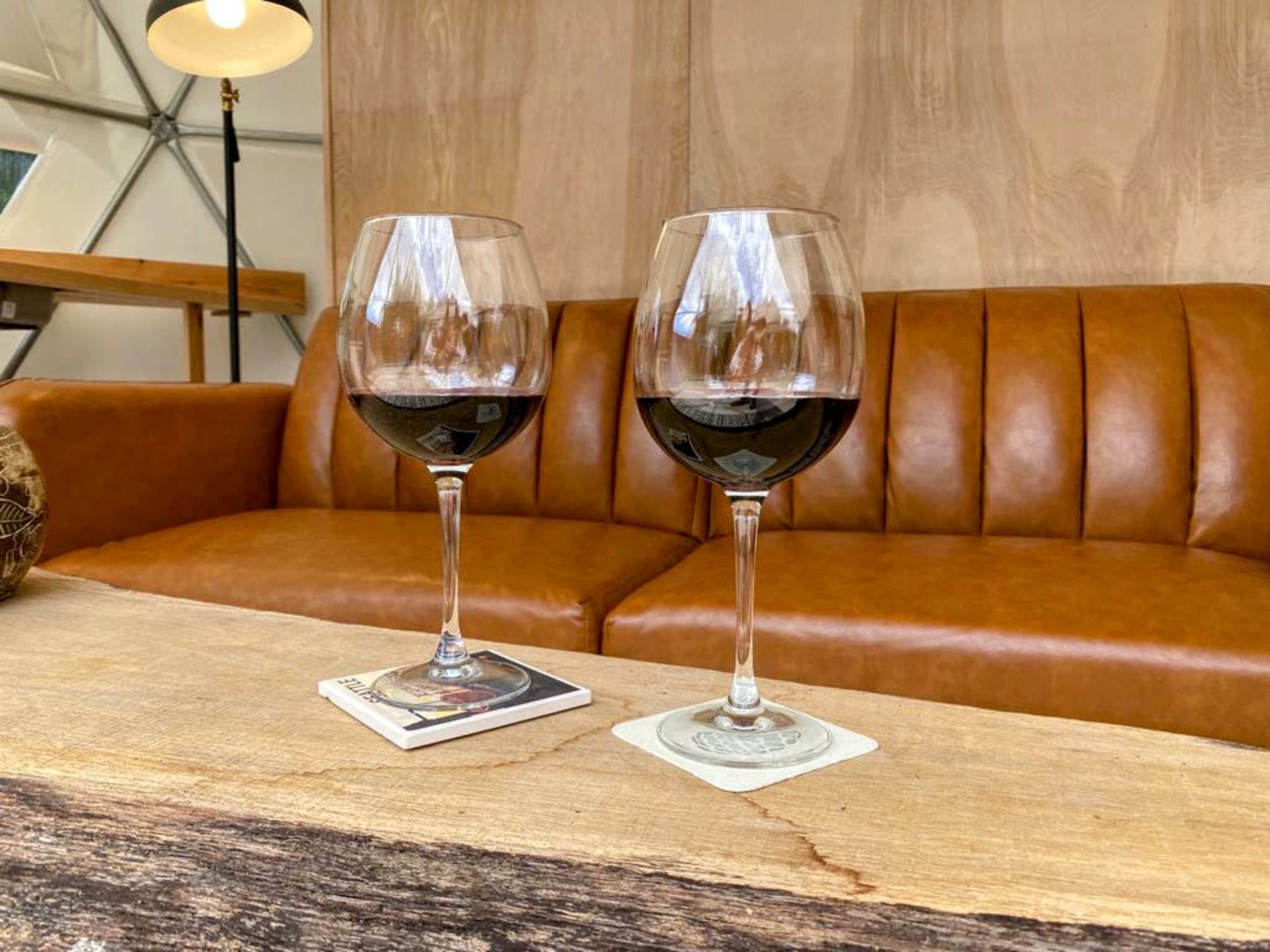 Two glasses of red wine are placed on coasters atop a wooden table in front of a brown leather couch.