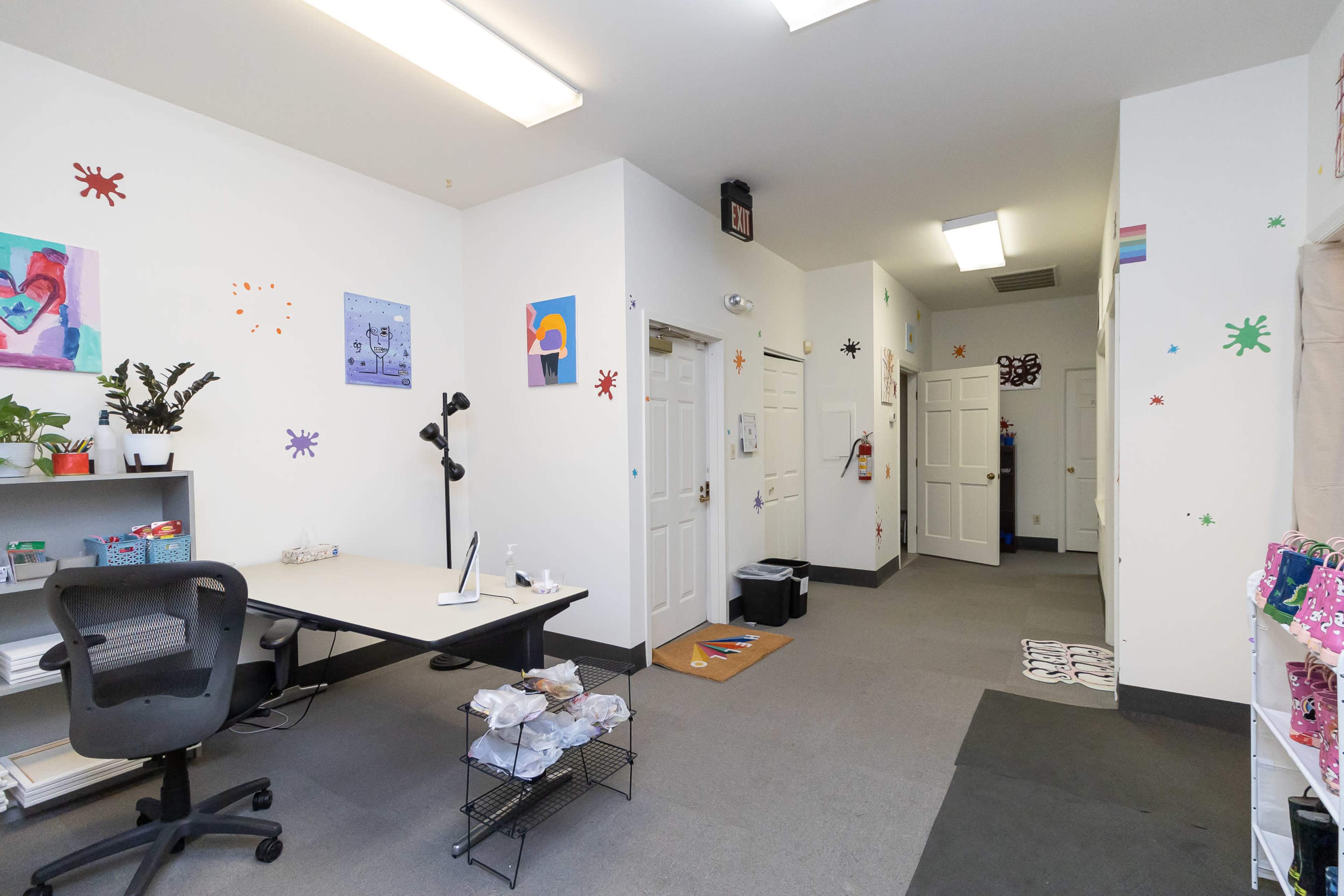 A brightly lit office space with a desk, chair, and decorative artwork on the walls, leading to a hallway with multiple doors.