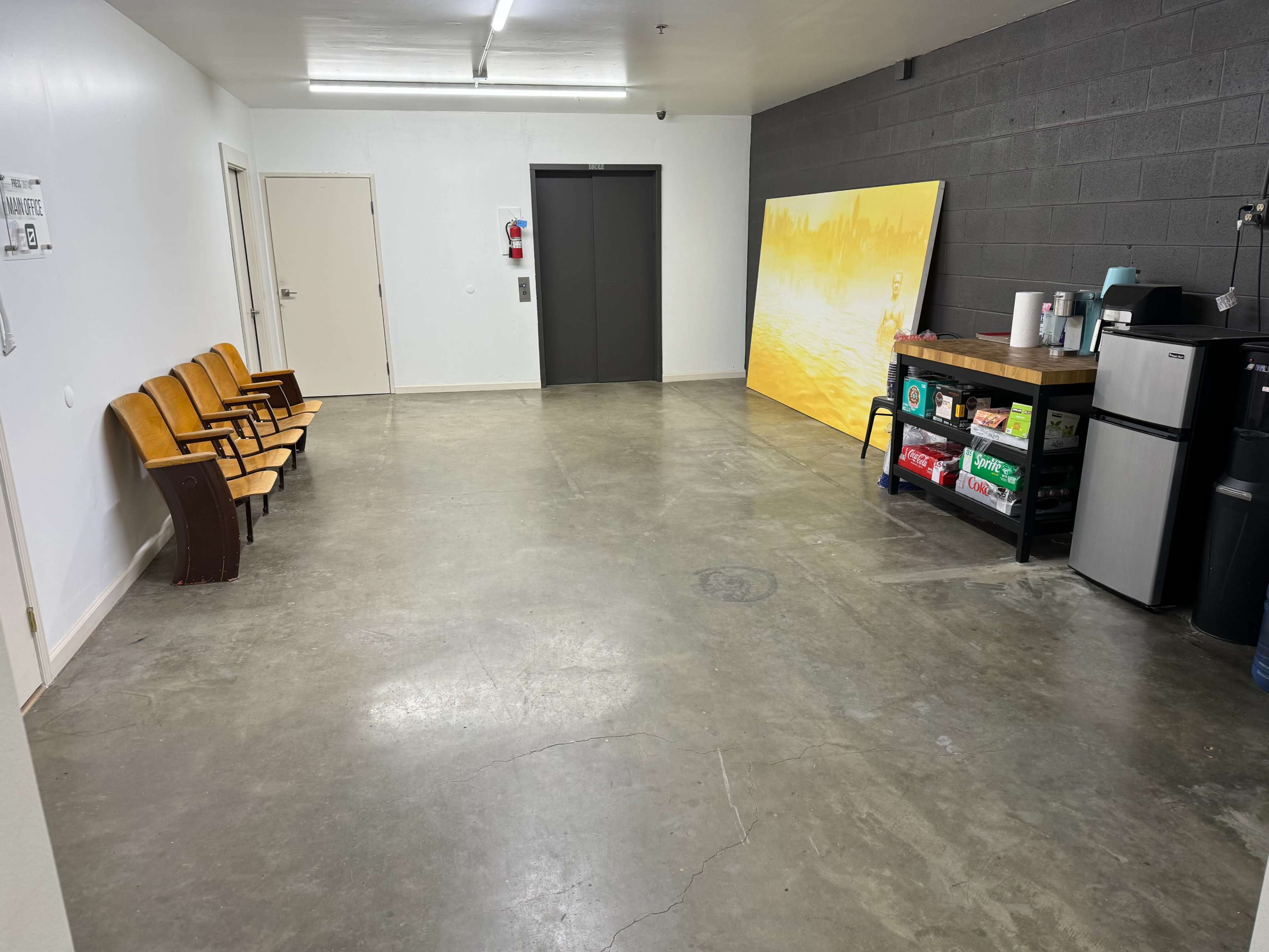 The image shows a spacious room with a concrete floor, several wooden chairs along one wall, an elevator door, and a kitchenette area with a table, fridge, and a large artwork on the wall.