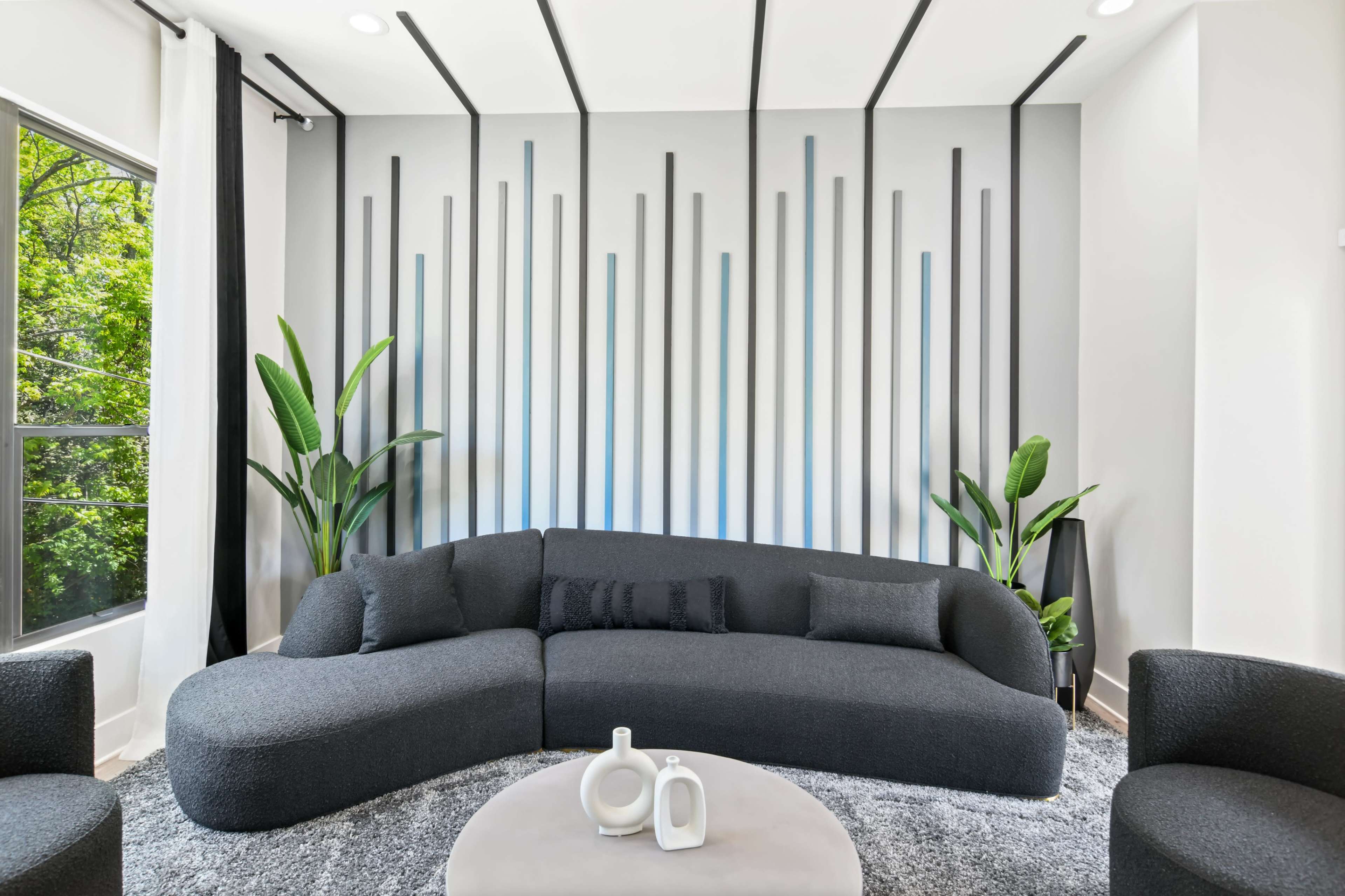A modern living room features a curved gray sofa, a round coffee table, and plants beside large windows.