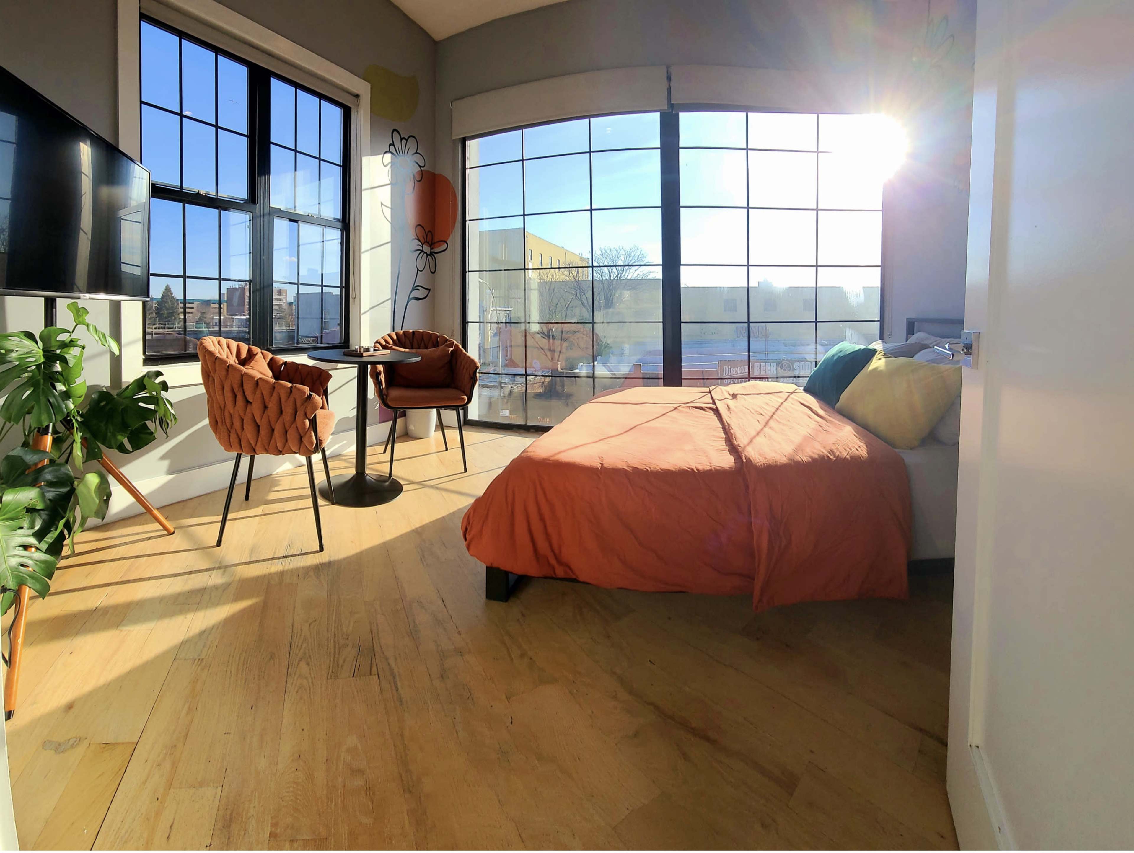 A brightly lit bedroom features a bed with an orange cover, two chairs at a small table, and large windows providing a view of the outdoors.