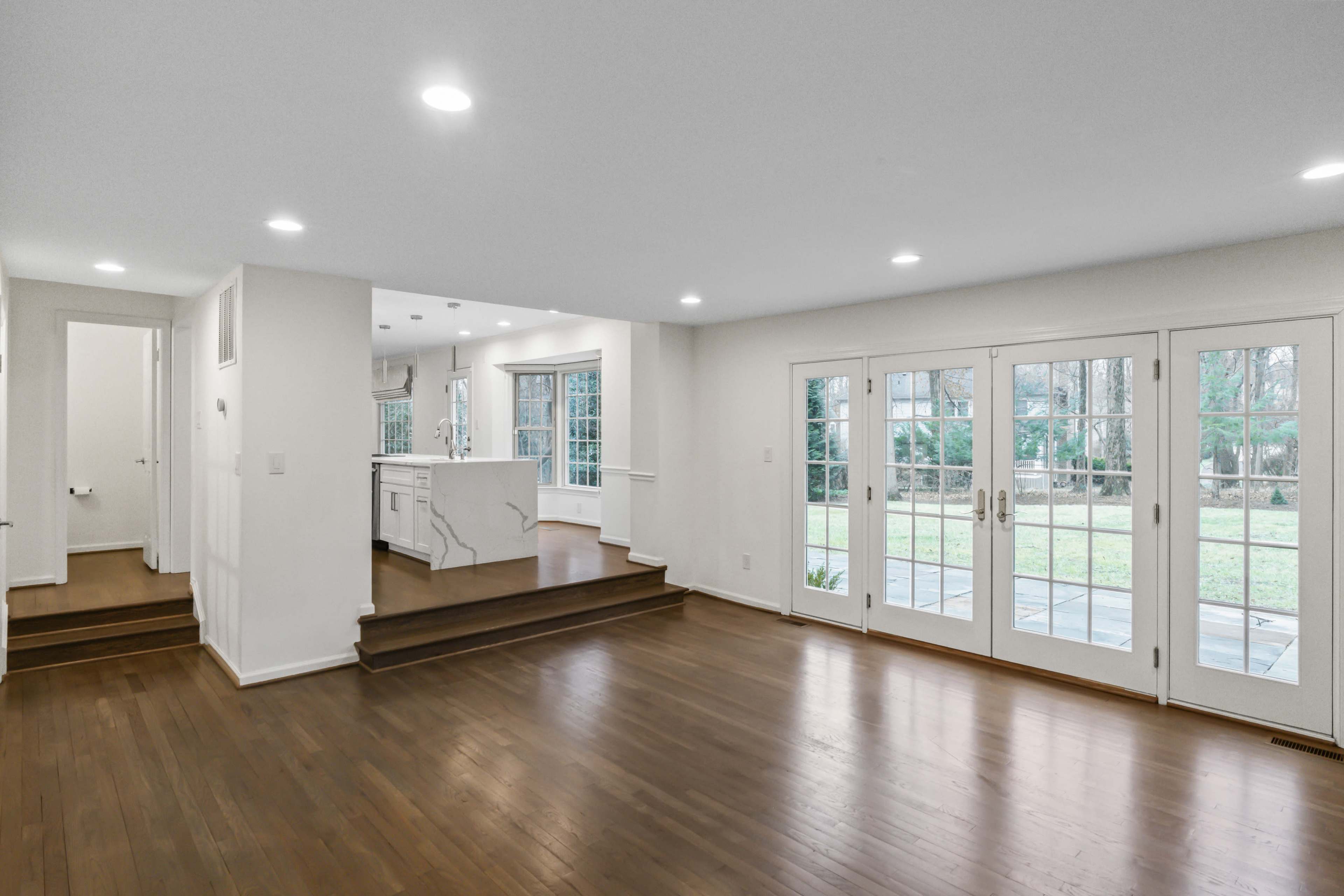 The image shows a spacious living area with hardwood floors, large windows, and access to a backyard through double glass doors.