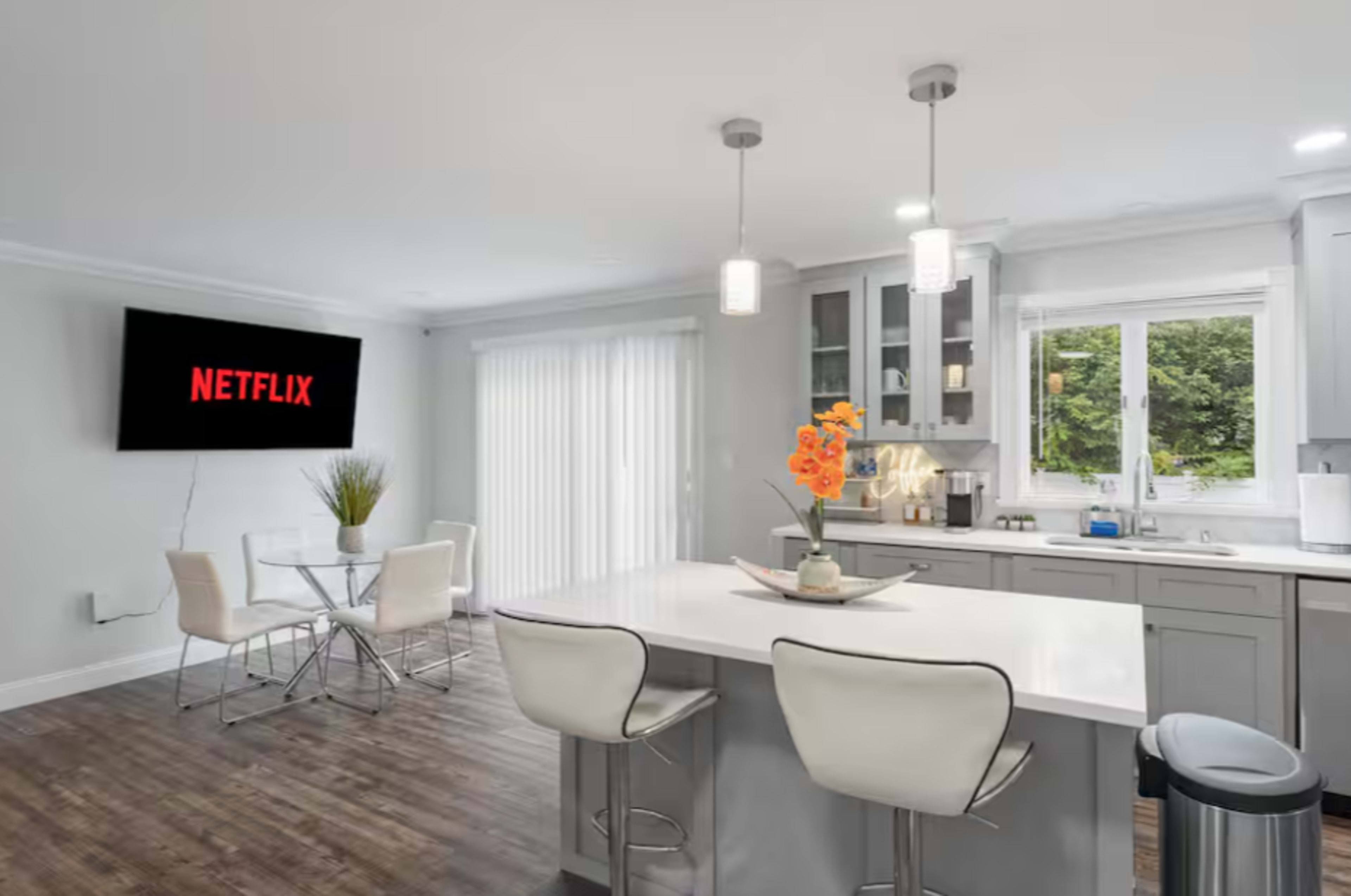 A modern kitchen features a central island with bar stools, a dining table, and a large screen displaying the Netflix logo on the wall.