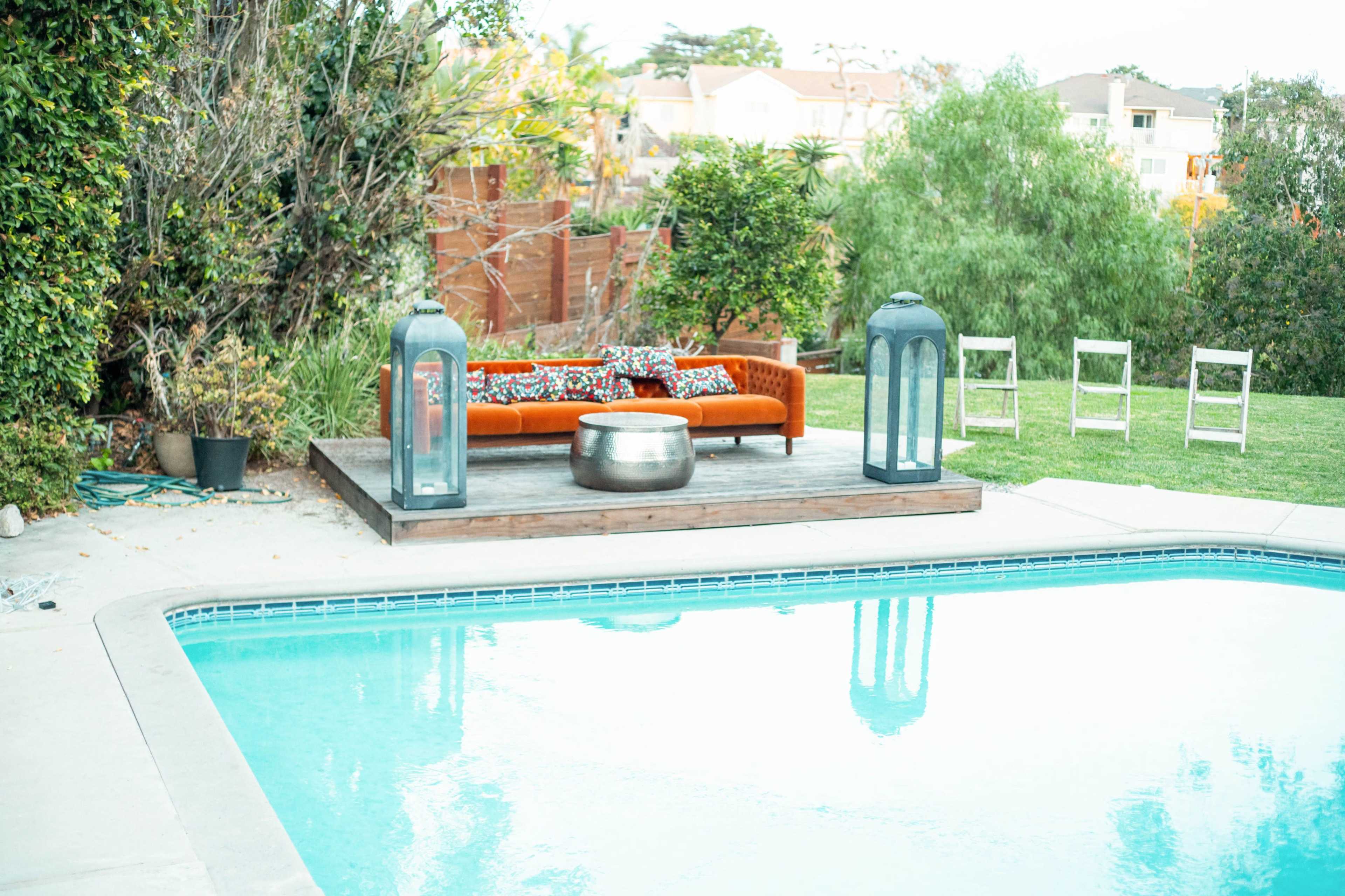 A swimming pool is surrounded by a wooden deck featuring a couch with decorative pillows, two lanterns, and a metal table, with chairs arranged nearby on the grass.