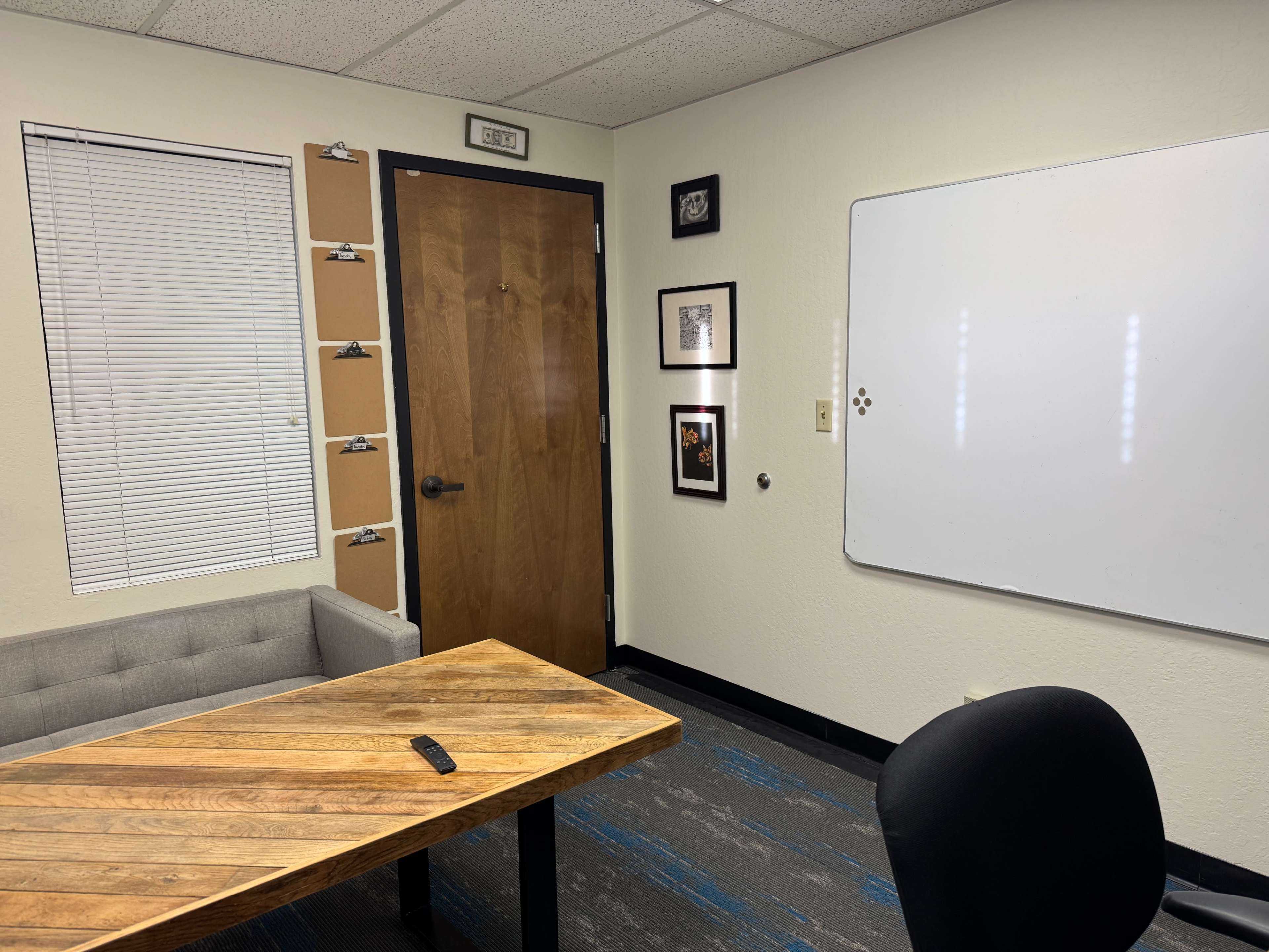 The image shows a small office with a wooden table, a gray couch, a whiteboard, and framed pictures on the walls.