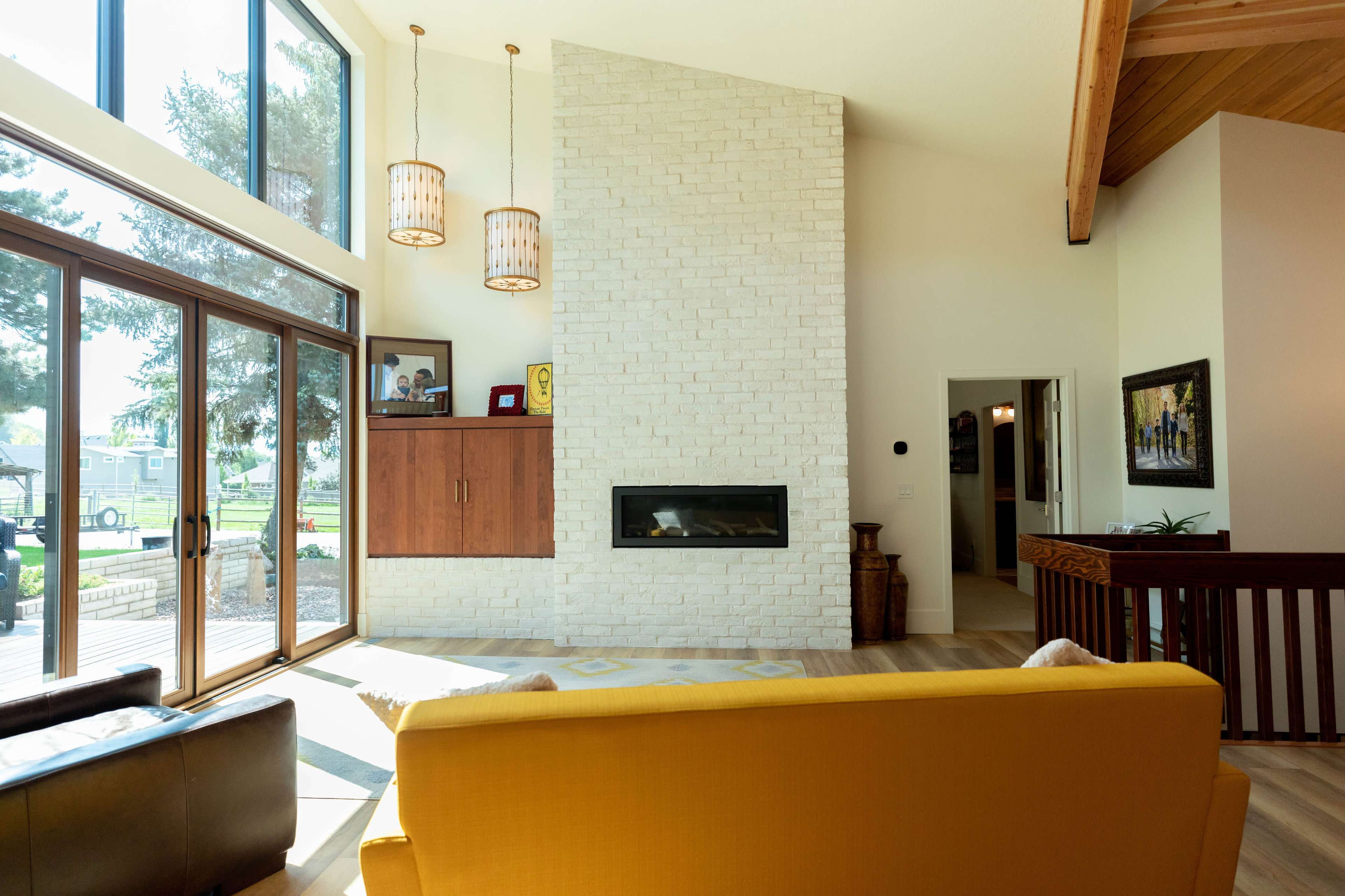 A modern living room features a yellow sofa facing a wall-mounted fireplace, large windows, and wooden ceiling beams.