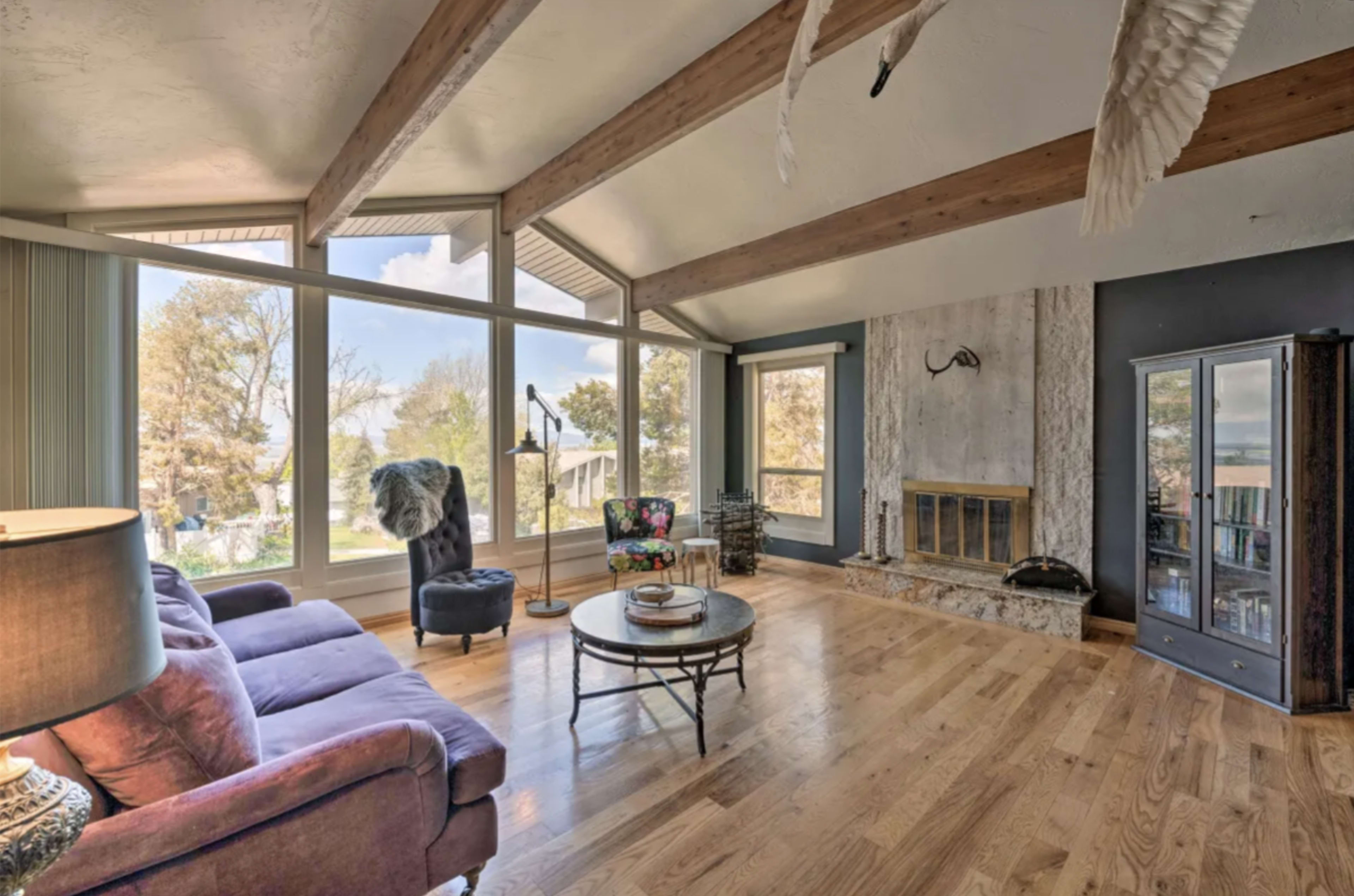The living room features large windows, wooden beams, and a stone fireplace, with a purple sofa and a round coffee table.