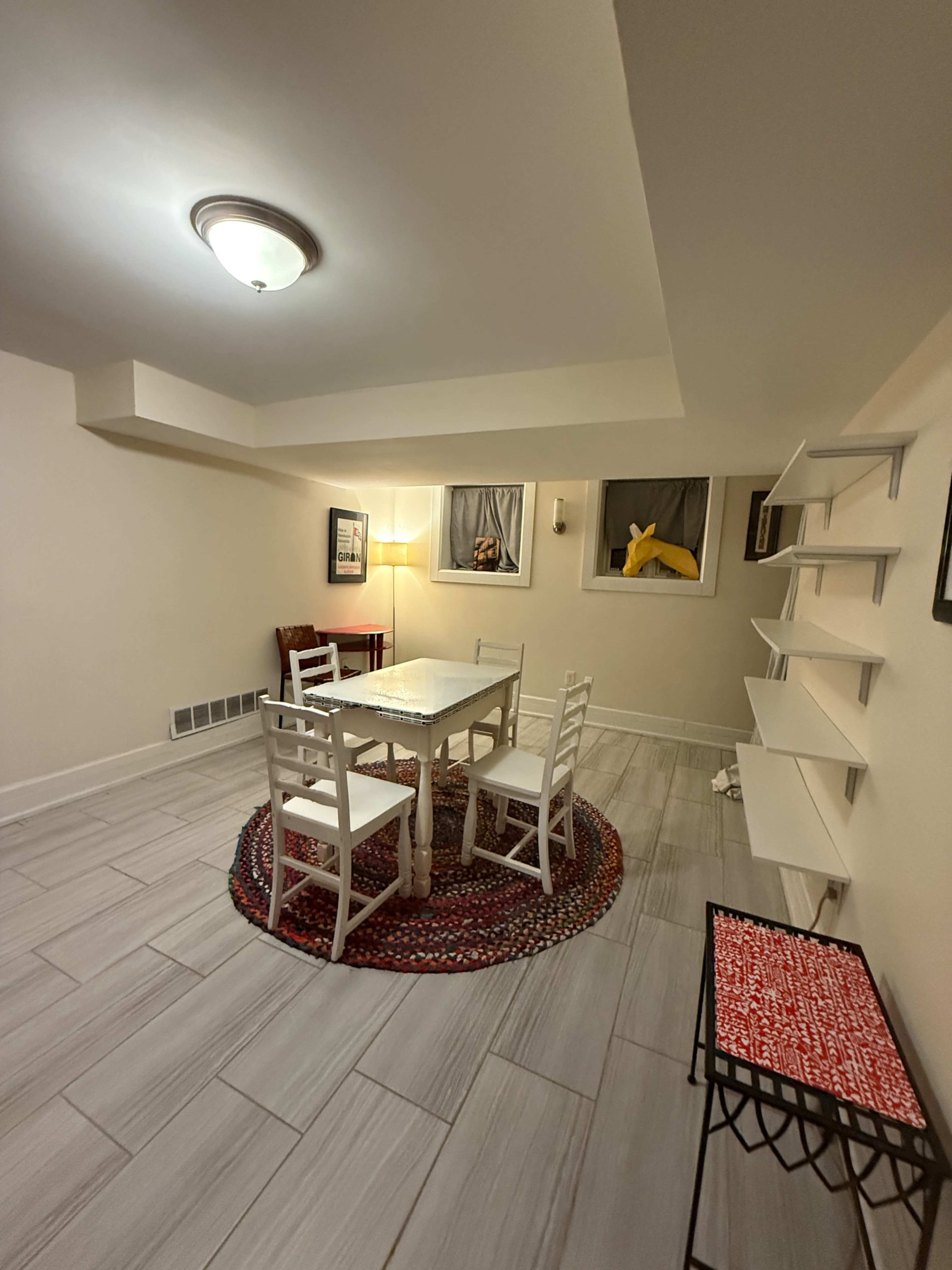 The image shows a small, well-lit basement room featuring a table surrounded by four white chairs, a circular rug, and shelves along the wall.