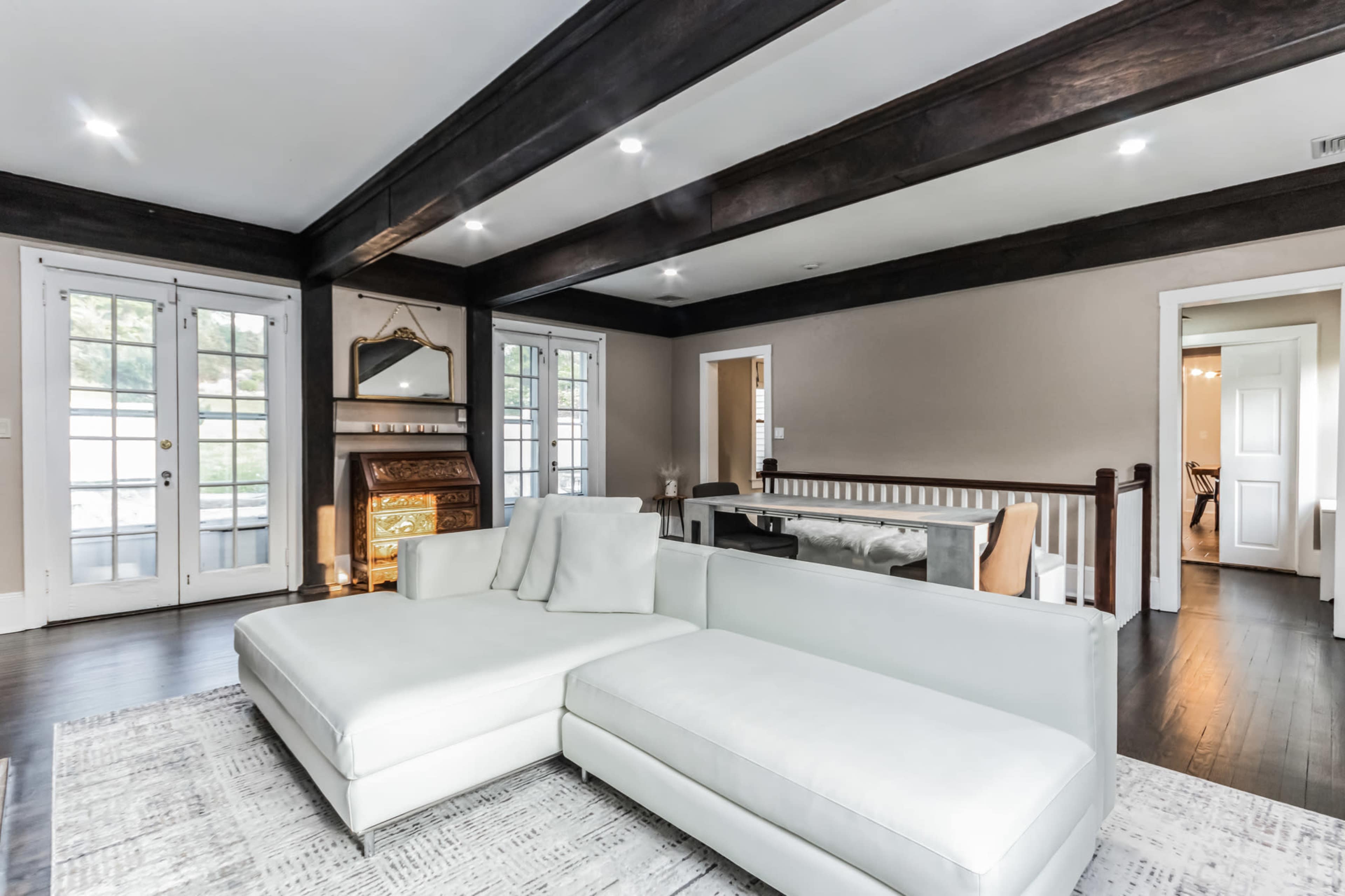 A modern living room features a white sectional sofa, wooden beams on the ceiling, and large windows offering natural light.