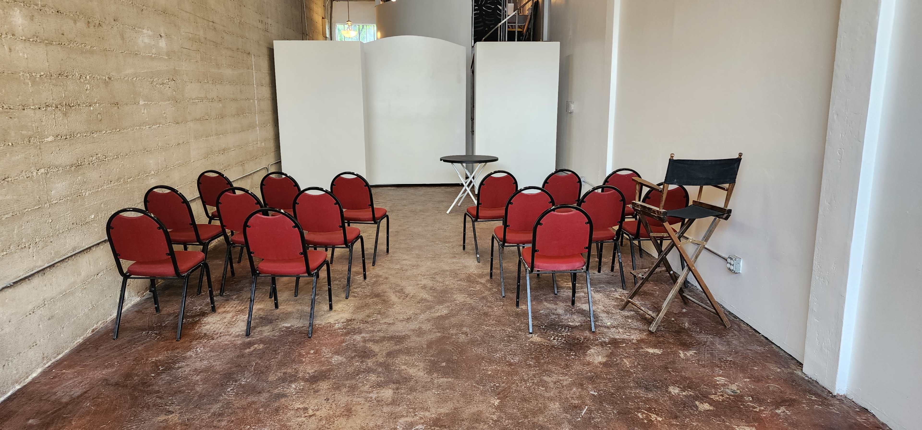 A room with a concrete wall featuring several rows of red chairs arranged around a round table, with a folding chair and a partition in the background.