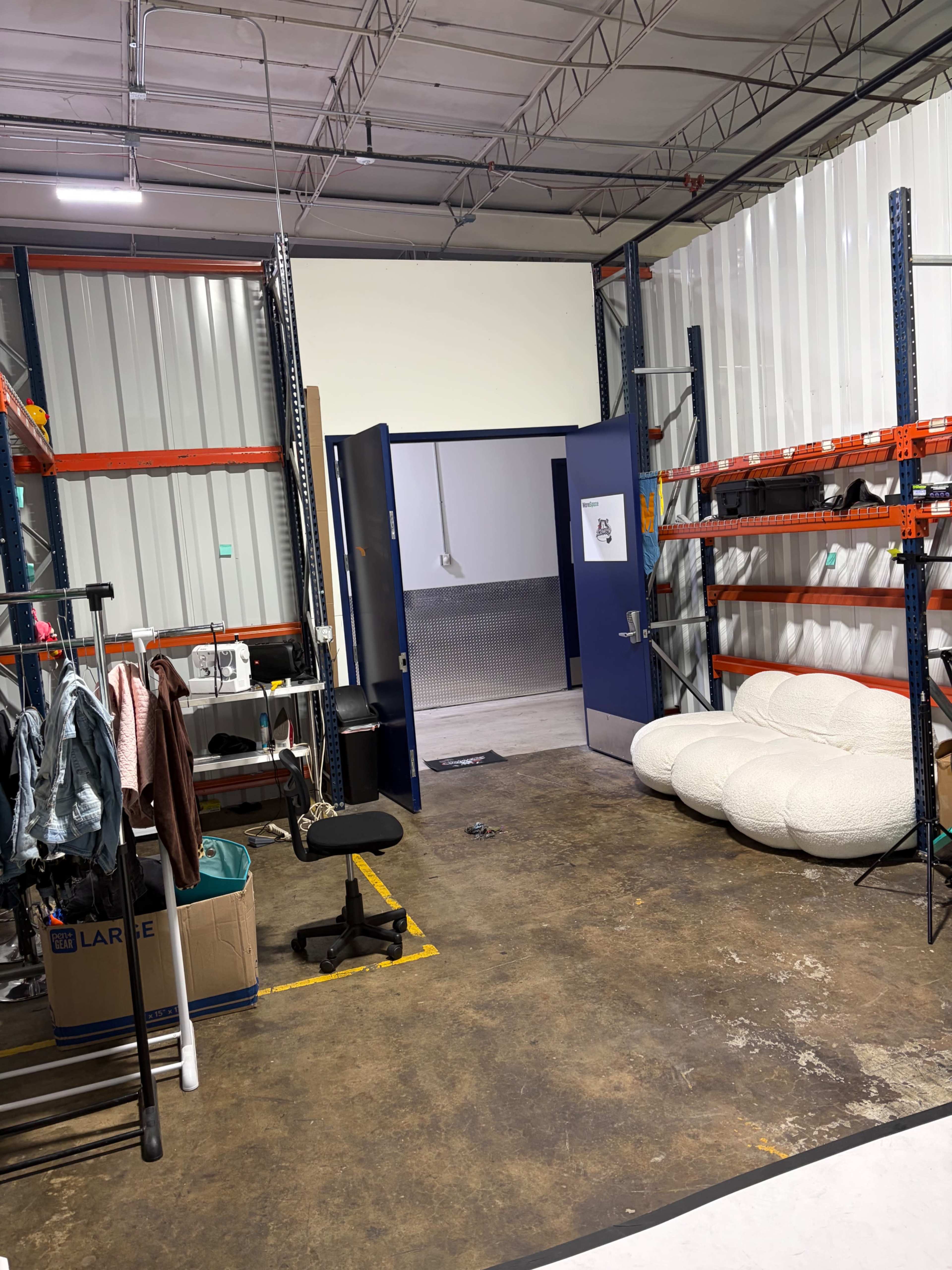 The image shows a storage area with metal shelves, a clothing rack, a chair, and a large, cushioned seating arrangement in the corner.