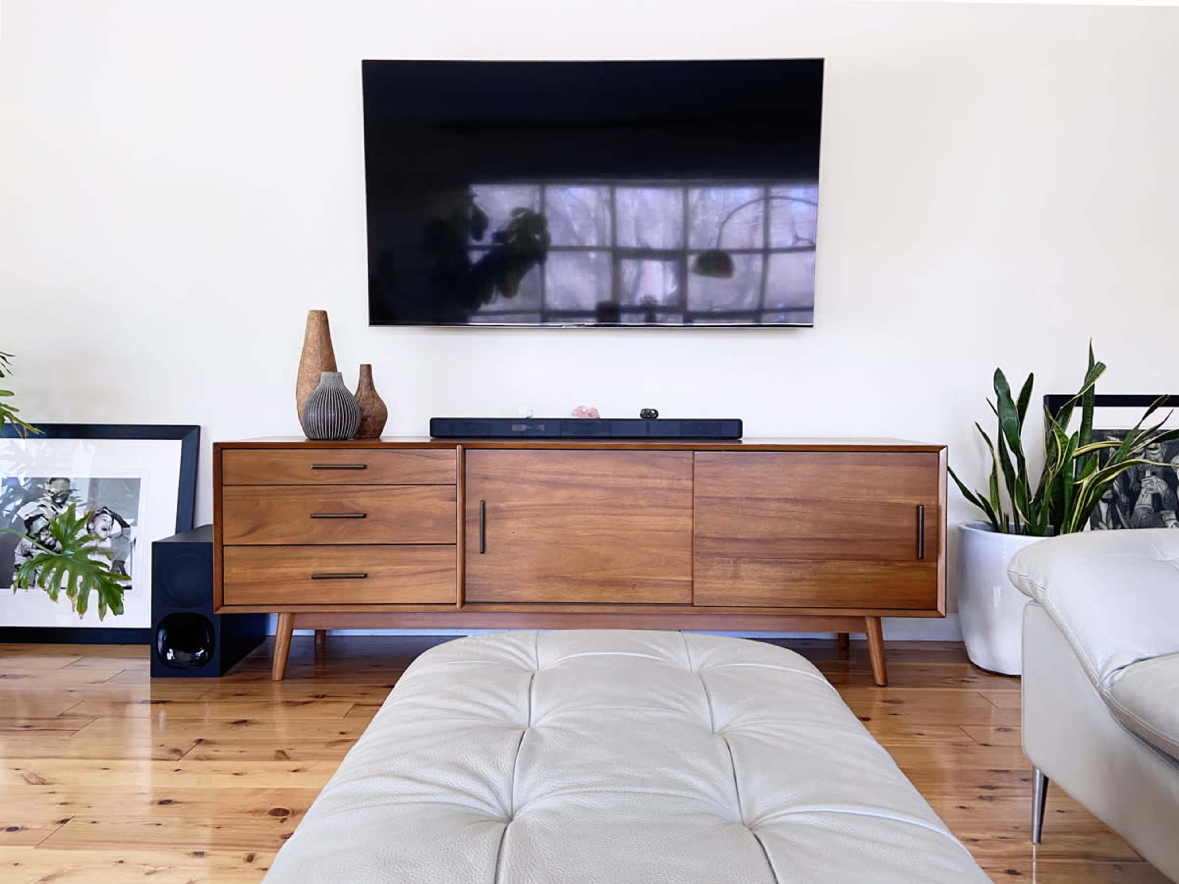 A wooden media console with drawers and decorative vases stands against a wall beneath a flat-screen TV, with potted plants and framed artwork nearby.