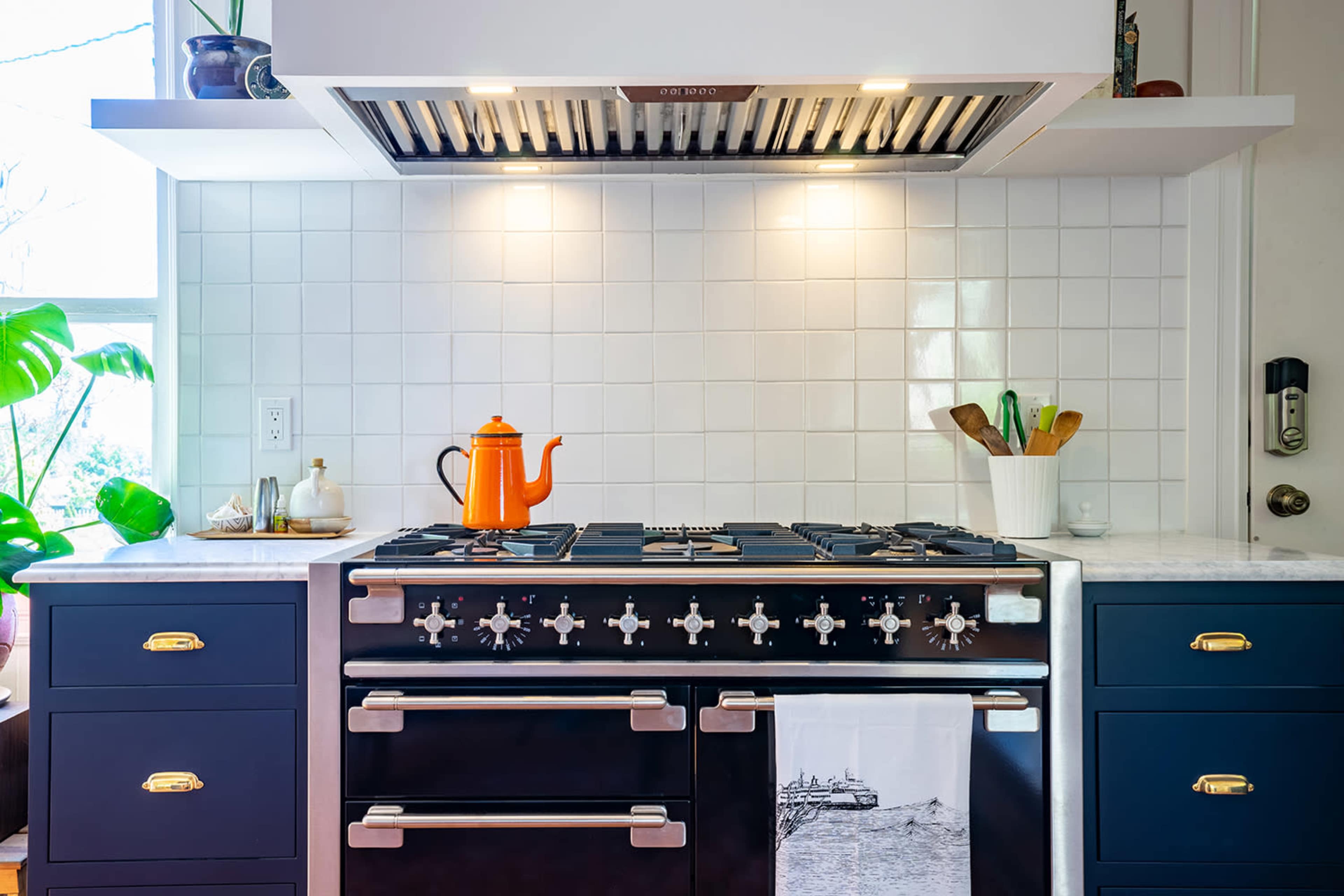 A modern kitchen with a gas stove, an orange kettle on the stove top, and a white tile backsplash.