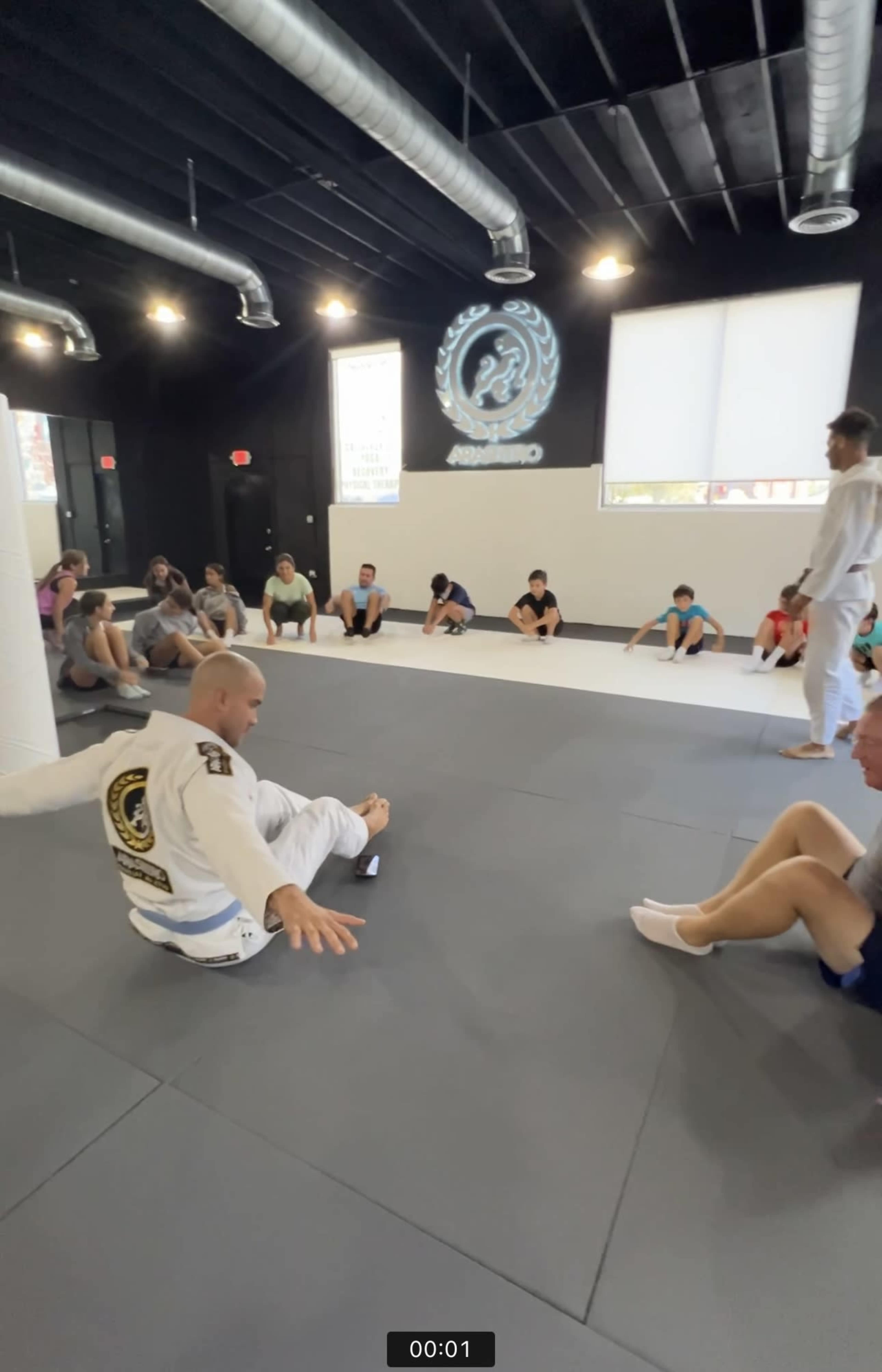 A group of people sits on a mat in a dojo while an instructor demonstrates a technique.