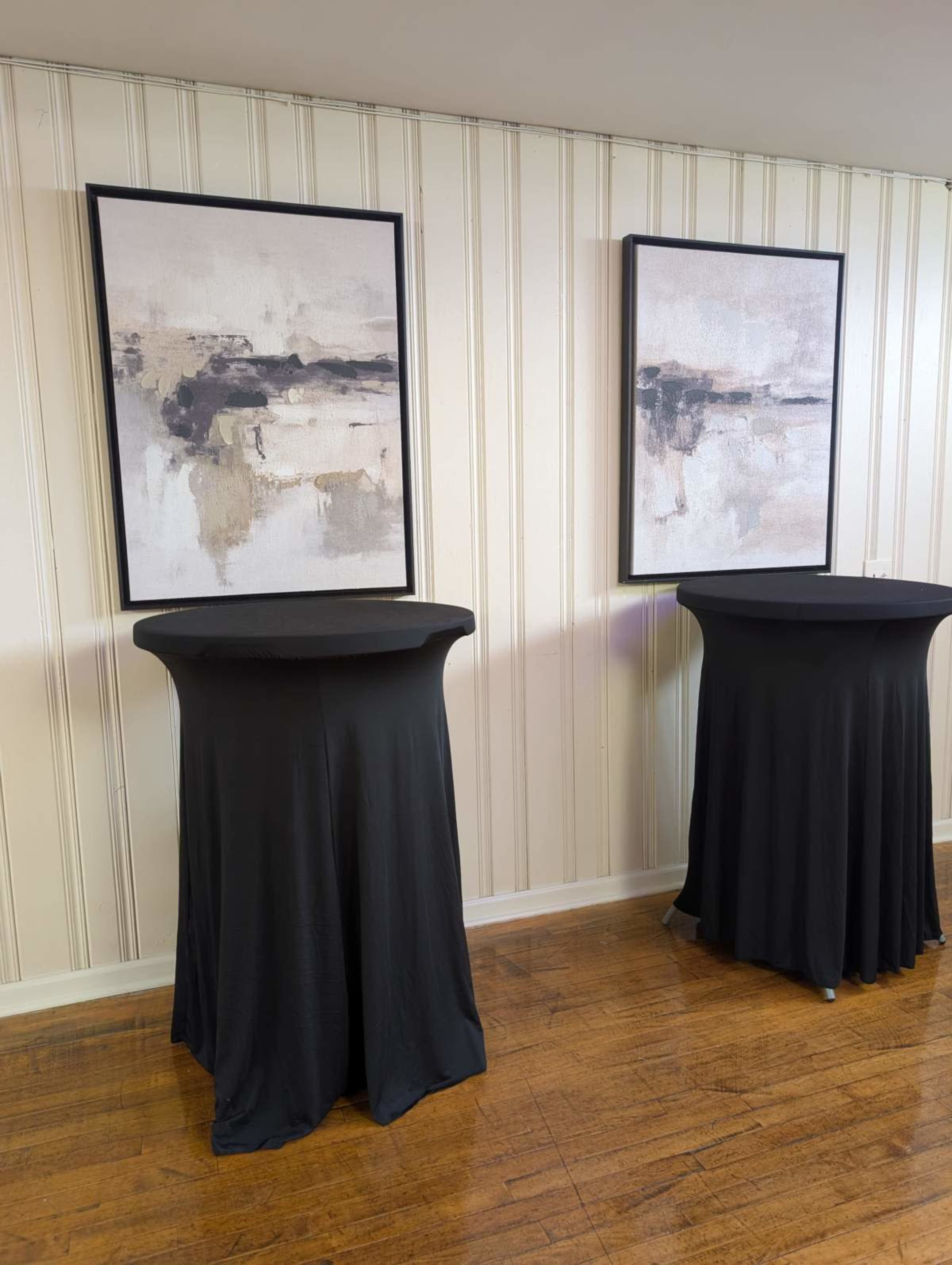 Two tall black tables covered with fitted tablecloths are positioned against a wall displaying two abstract paintings.