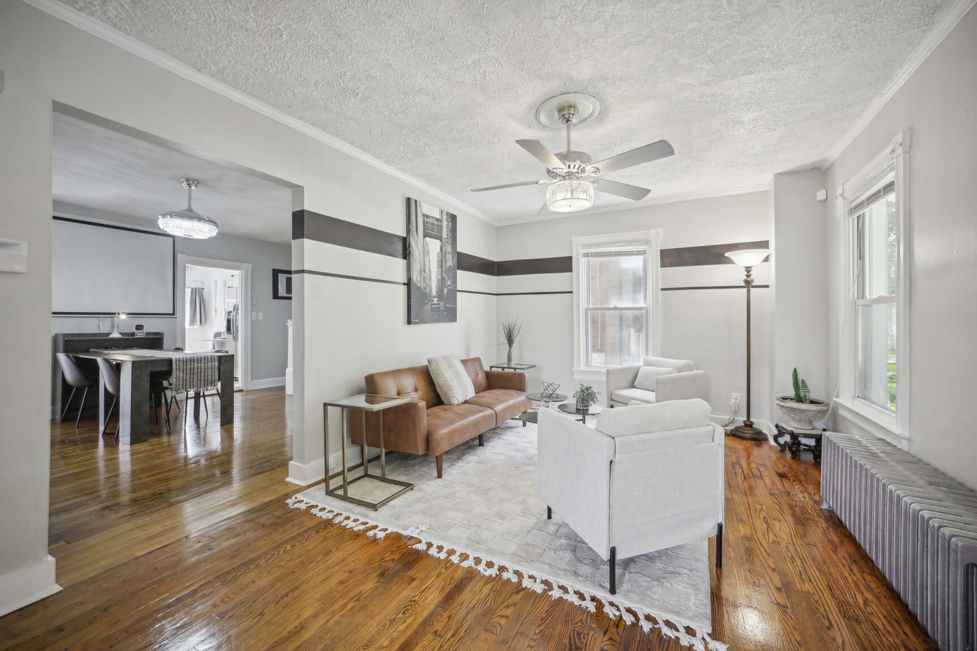 A living room with hardwood floors, a brown leather sofa, white armchairs, and a ceiling fan, leading into a dining area visible beyond.