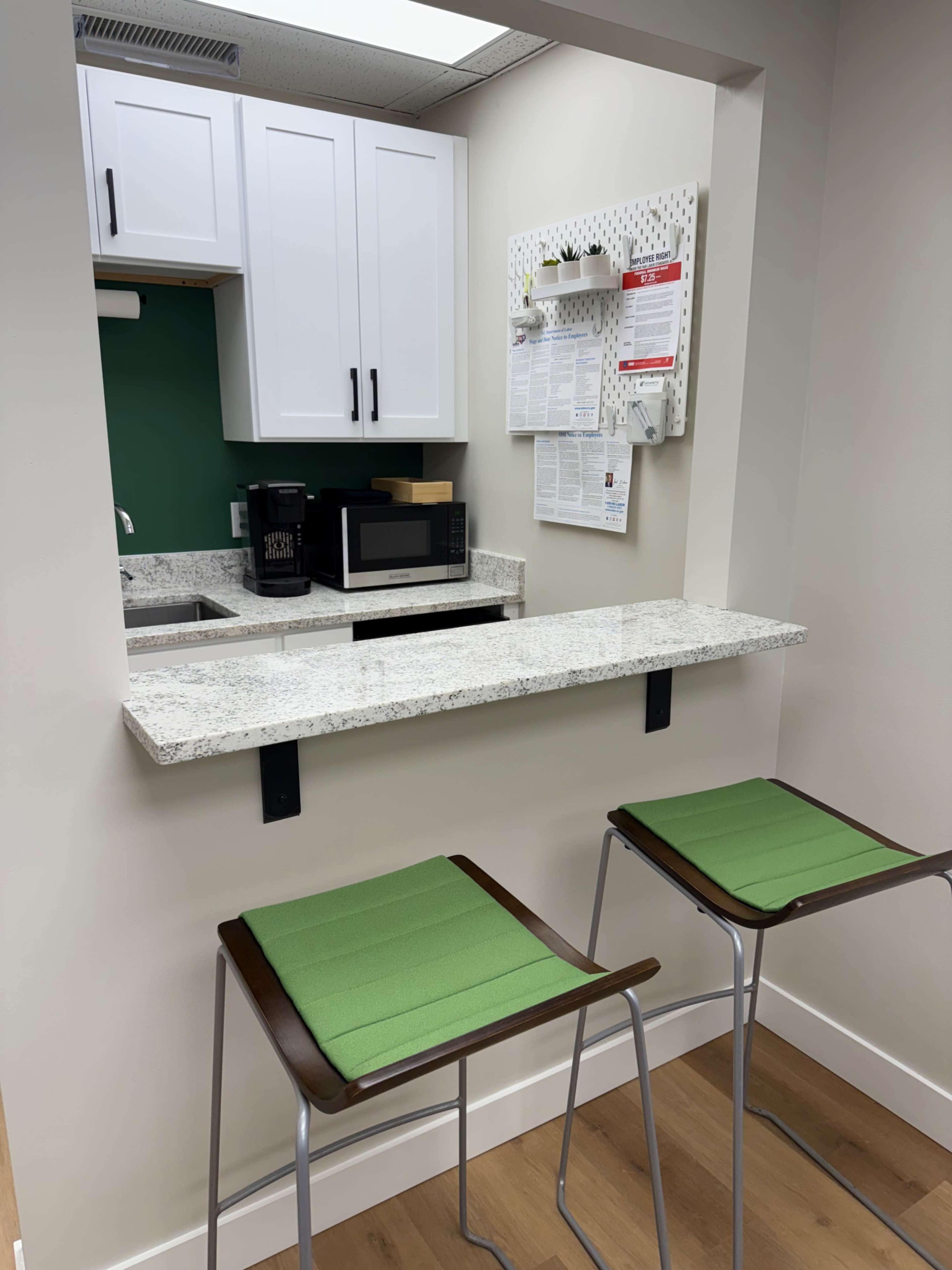 The image shows a small kitchen area featuring a countertop with two green stools, a microwave, and a calendar mounted on the wall.