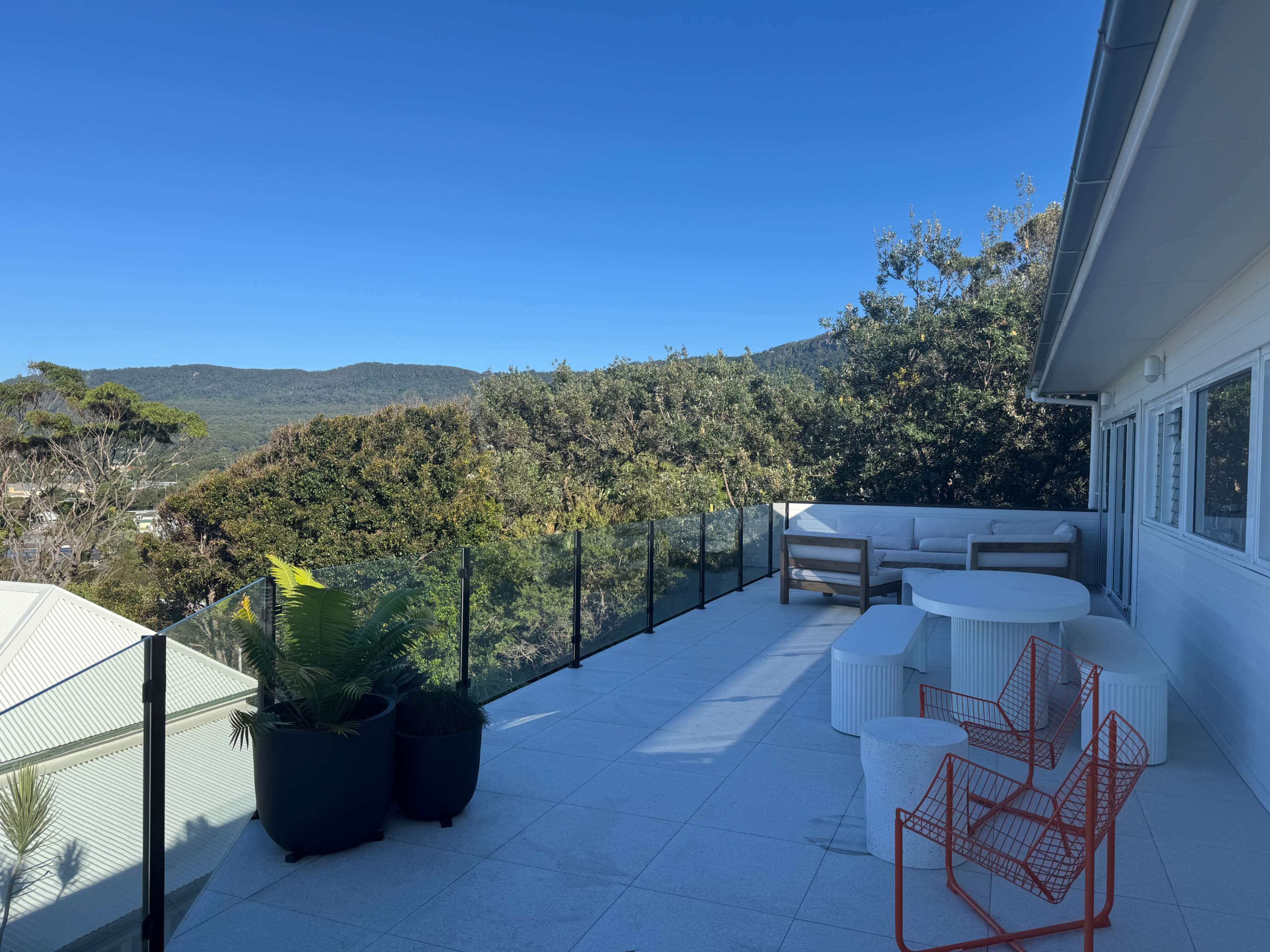 A spacious rooftop terrace features white seating, circular tables, and vibrant orange chairs, overlooking a green landscape and blue sky.