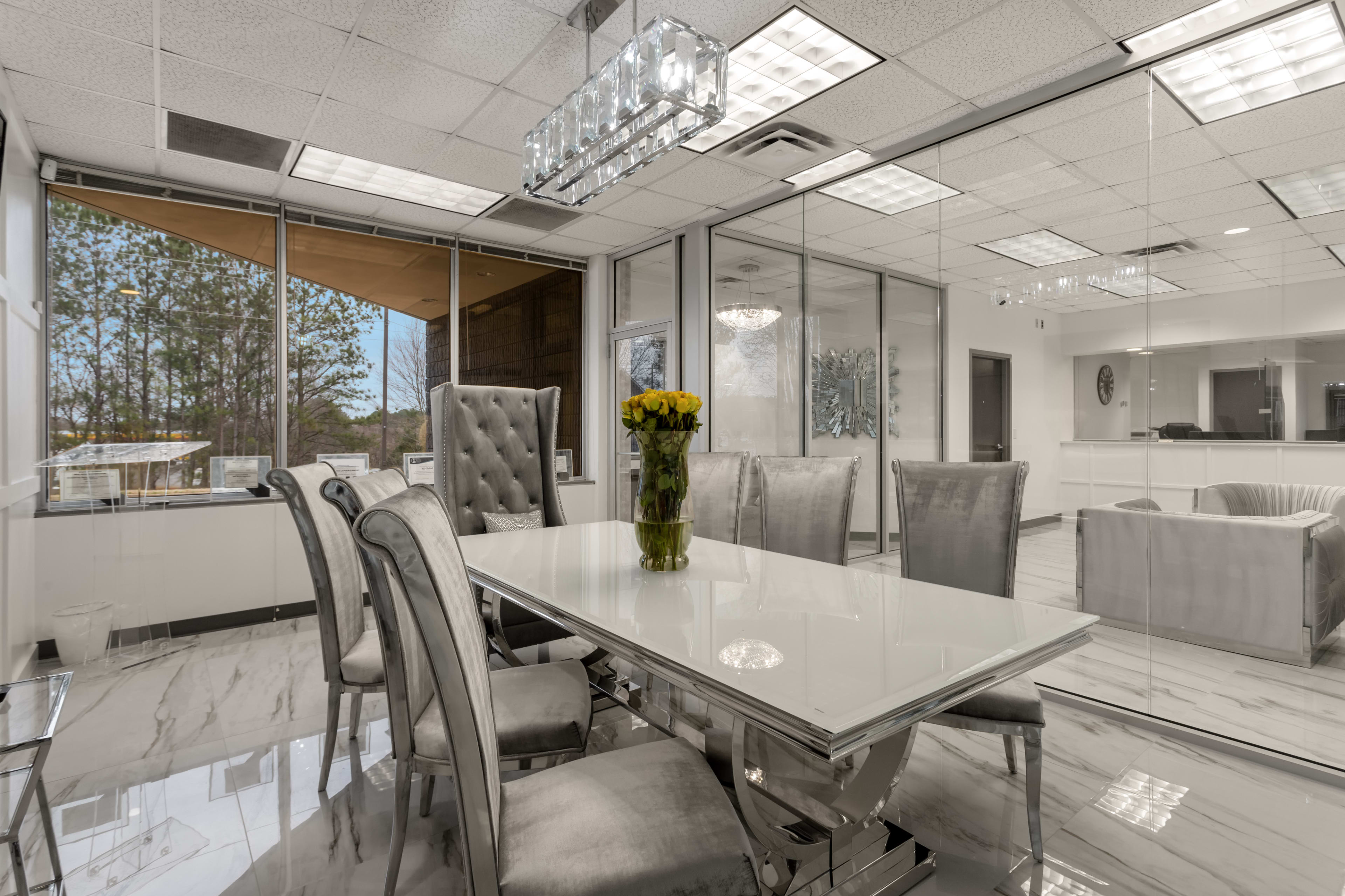 A modern glass-walled dining area features a long white table surrounded by upholstered chairs and a floral centerpiece.
