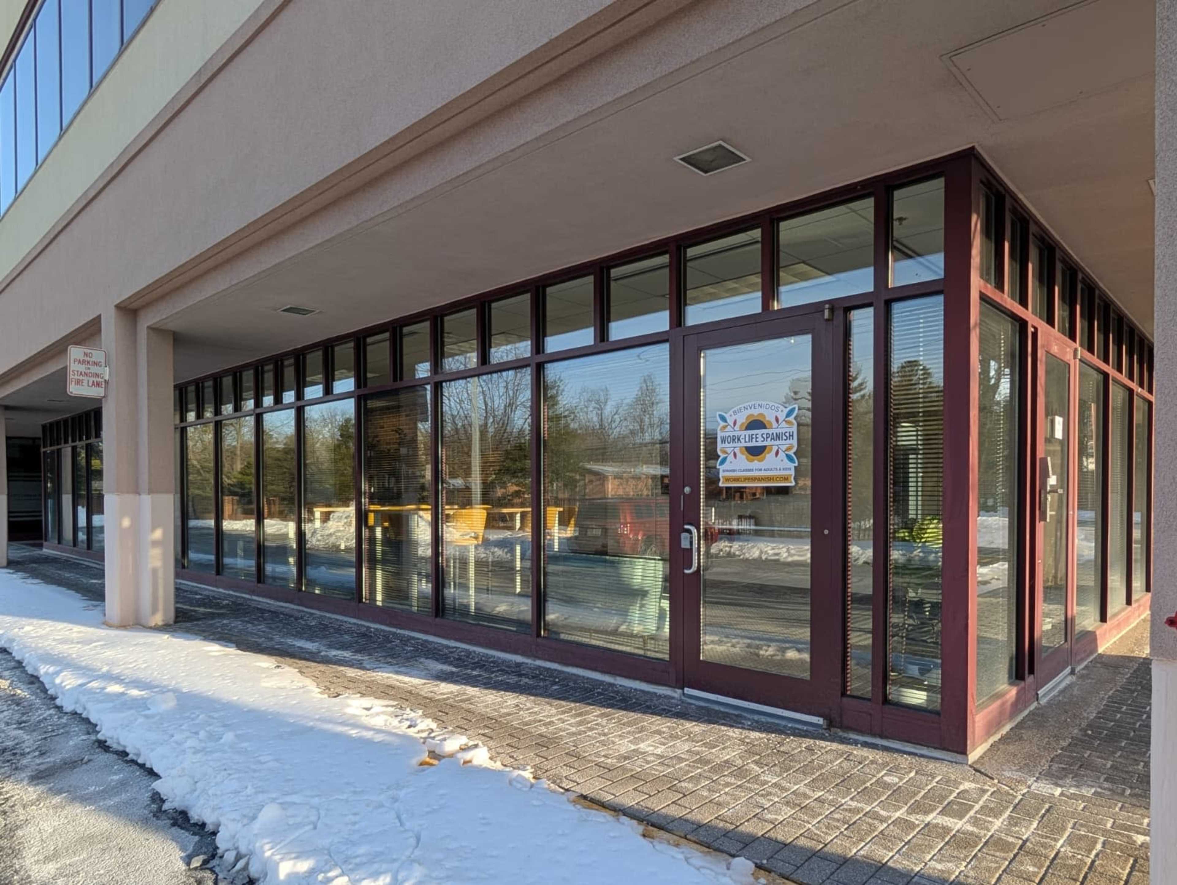 The image shows a storefront with large windows and a glass door, situated in a building with a concrete walkway and snow on the ground.