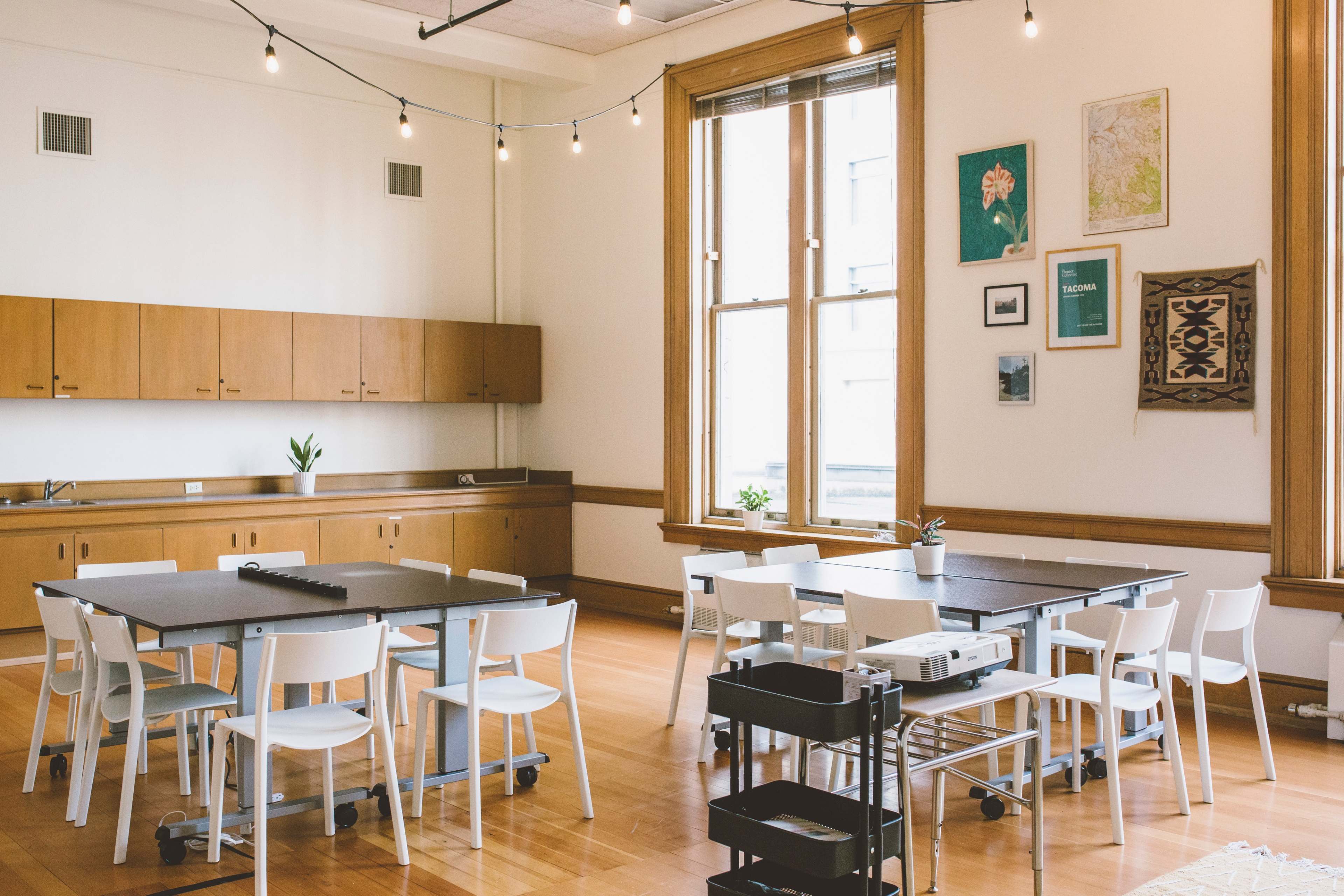 A well-lit, modern classroom or meeting space with several tables and chairs, cabinetry along one wall, and decorative artwork on the walls.