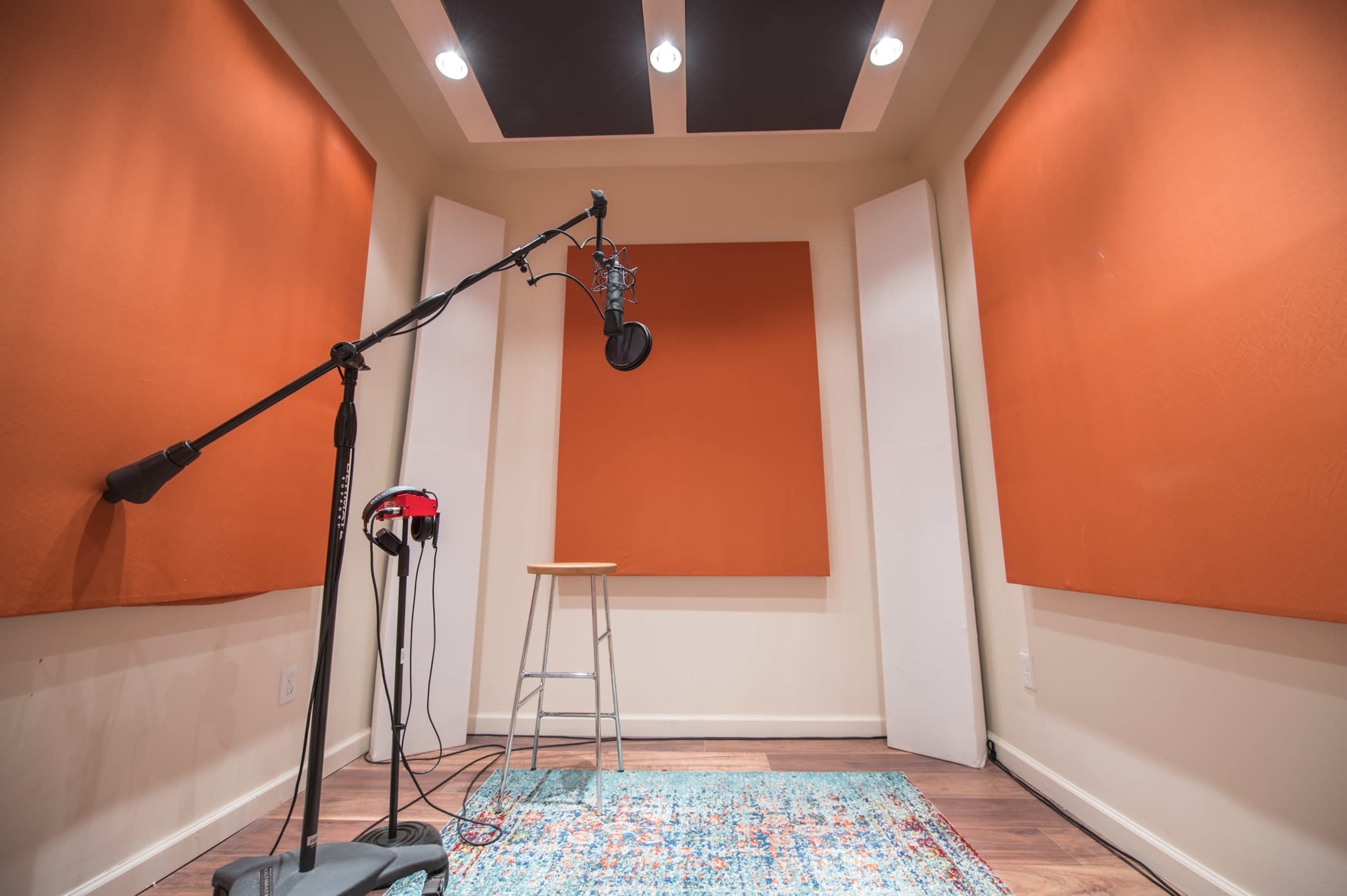 A recording studio booth features orange soundproofing panels on the walls, a microphone on a stand, a stool, and a rug on the floor.
