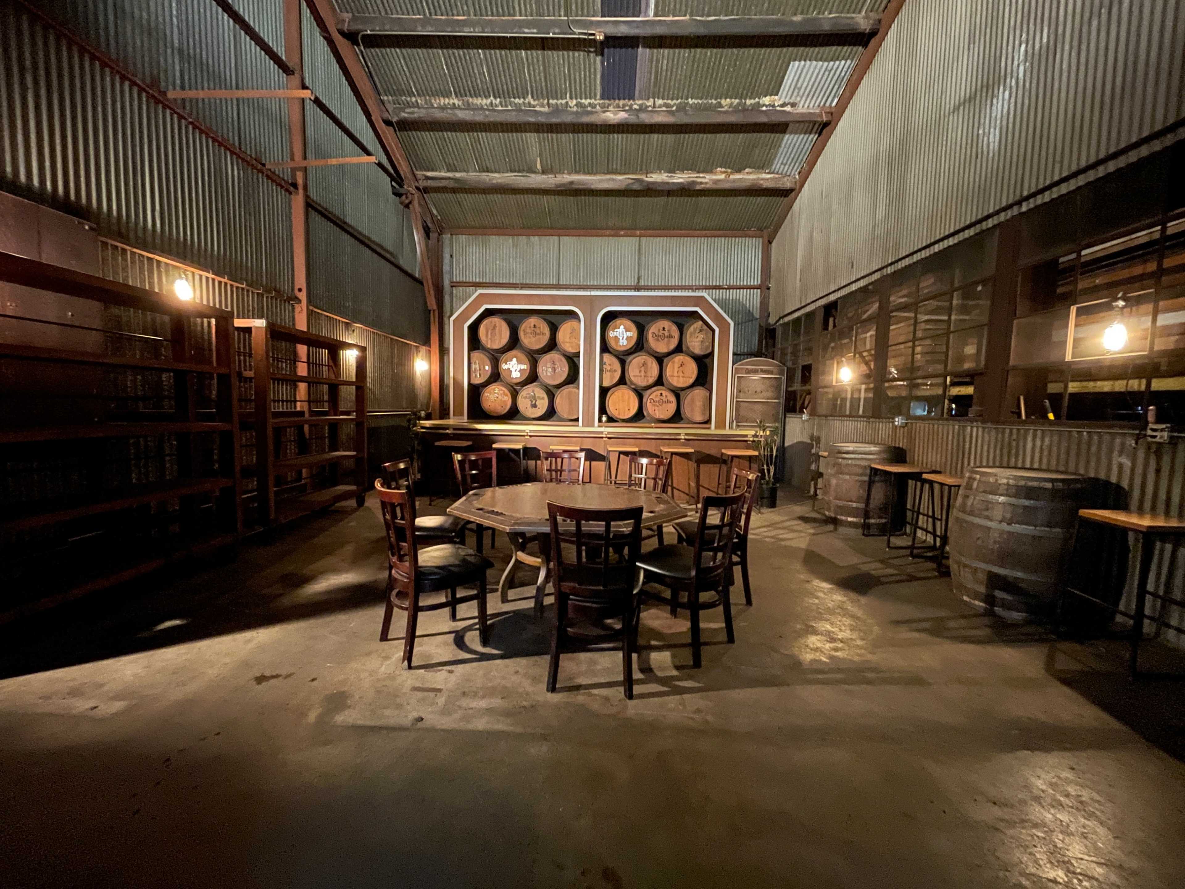 A dimly lit room features a wooden table surrounded by chairs, with shelves and barrels against the corrugated metal walls.