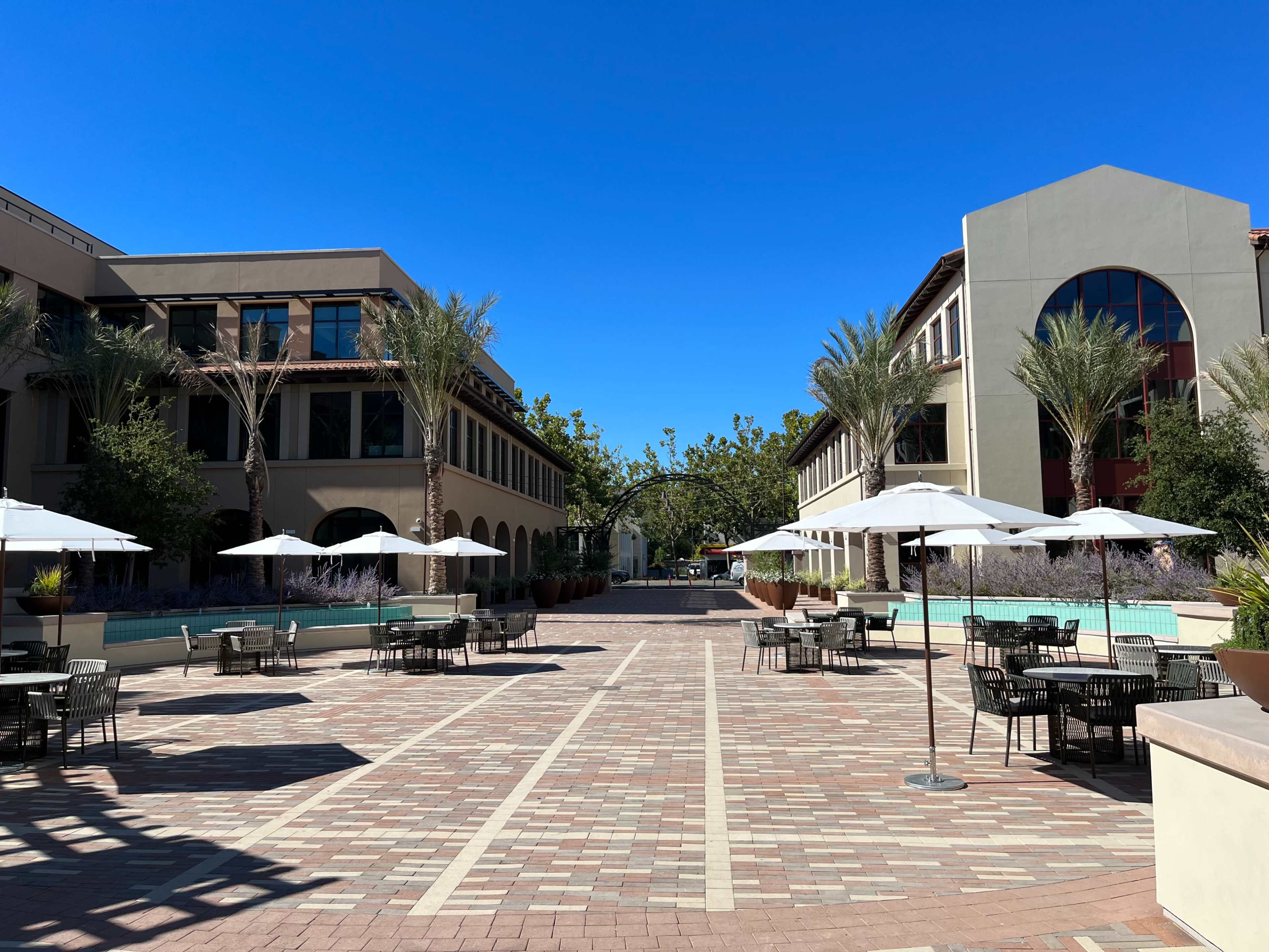 A brick plaza is flanked by two buildings, featuring several tables with umbrellas and palm trees.