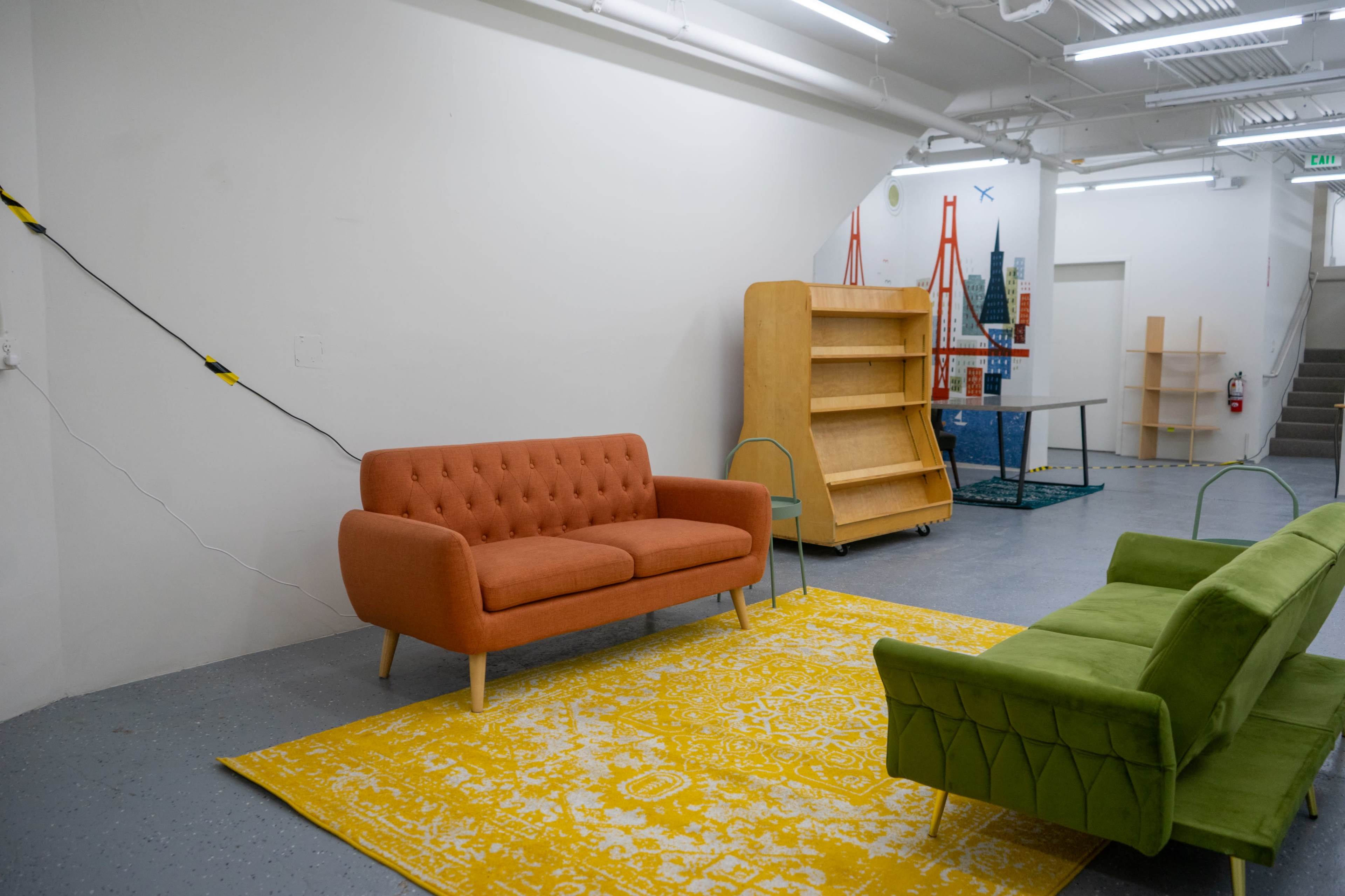 The image shows a minimalist room with an orange sofa, a green sofa, a yellow patterned rug, and wooden shelves against the wall.