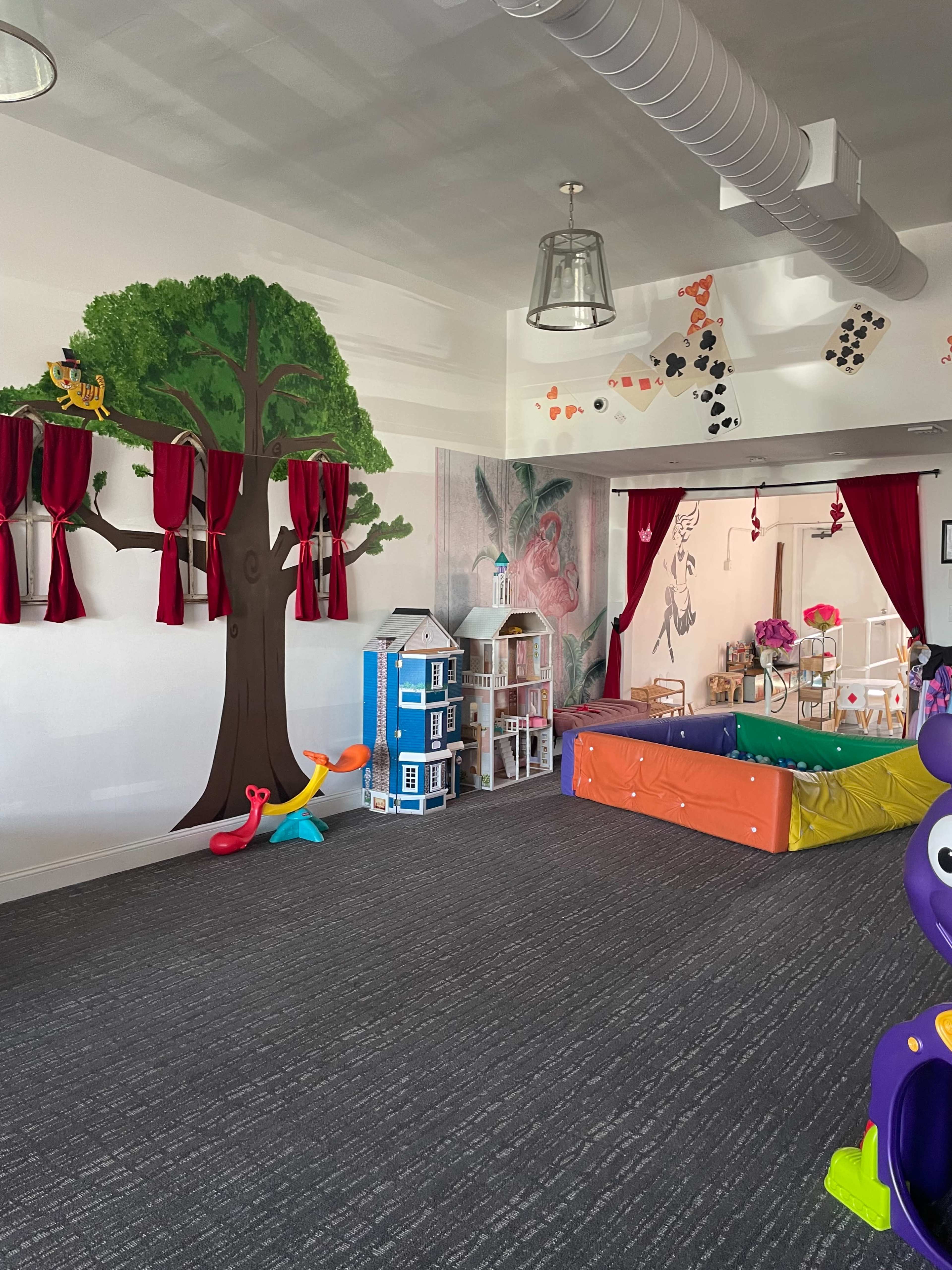 The image shows a children's playroom with a large tree mural on one wall, colorful play equipment, and a blue dollhouse shelf.