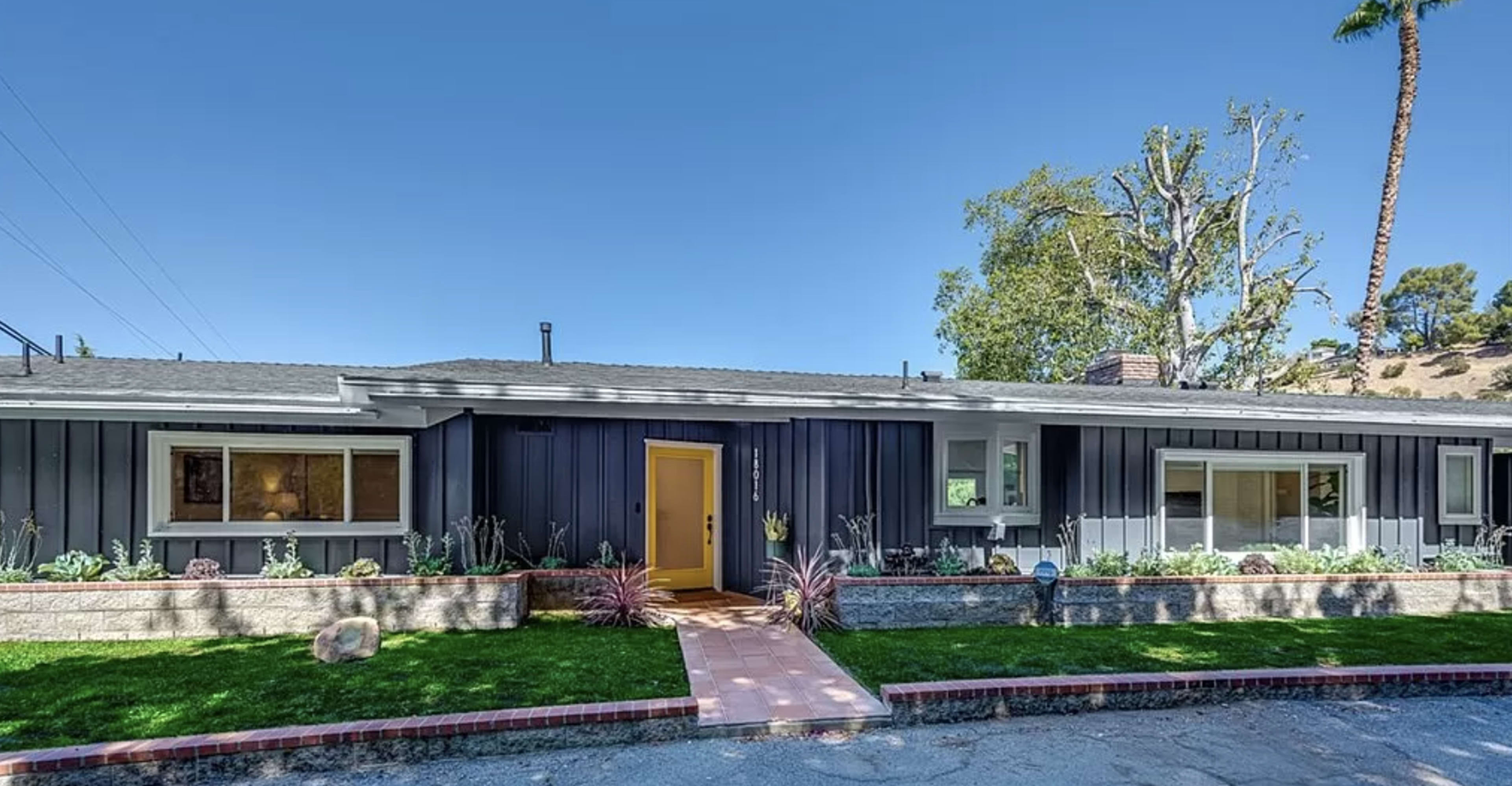 A single-story, dark-colored house with a yellow front door and a landscaped yard featuring plants and a stone pathway.