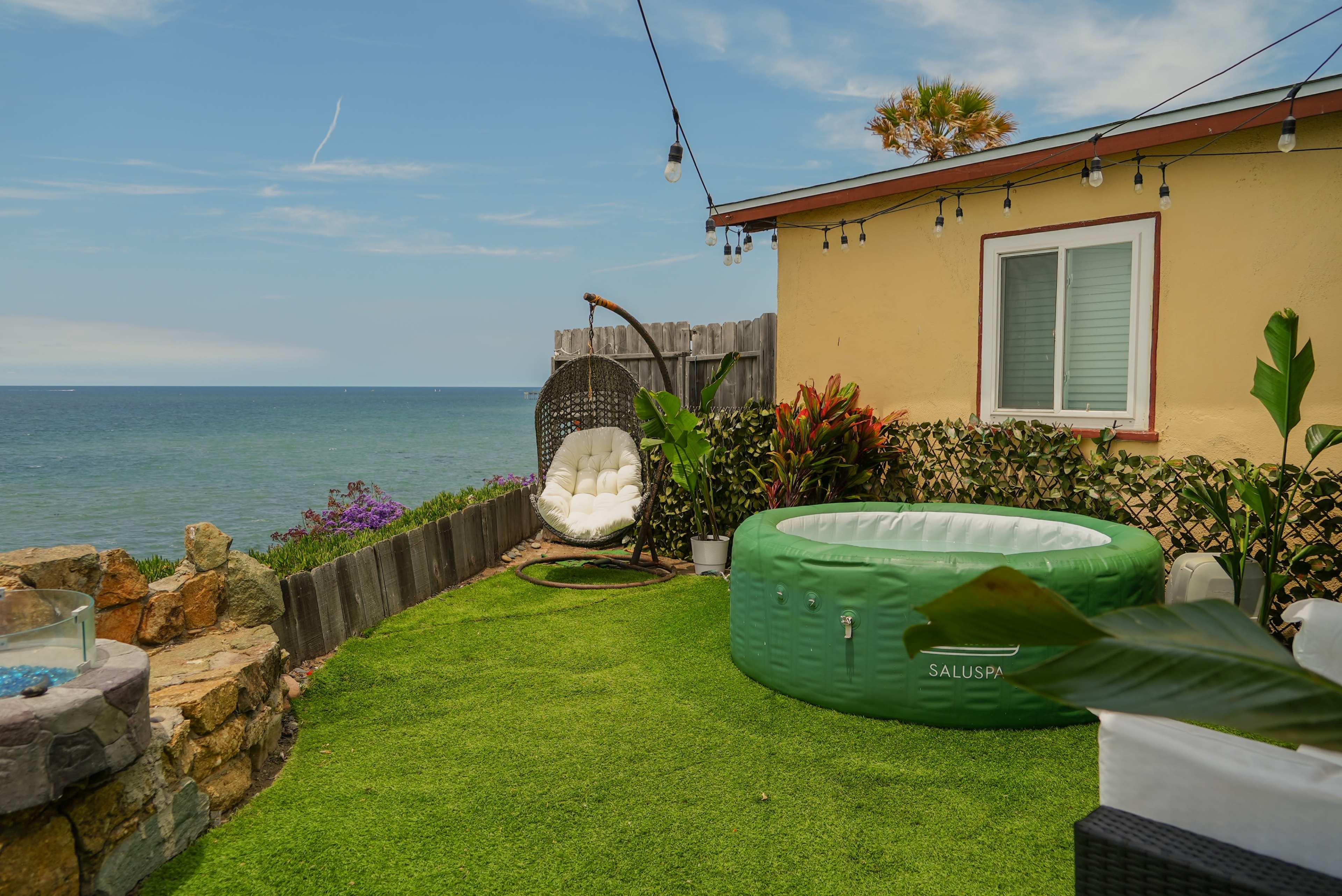 A green inflatable hot tub is situated on a grassy area near the ocean, alongside a hanging chair and colorful plants, with a house in the background.