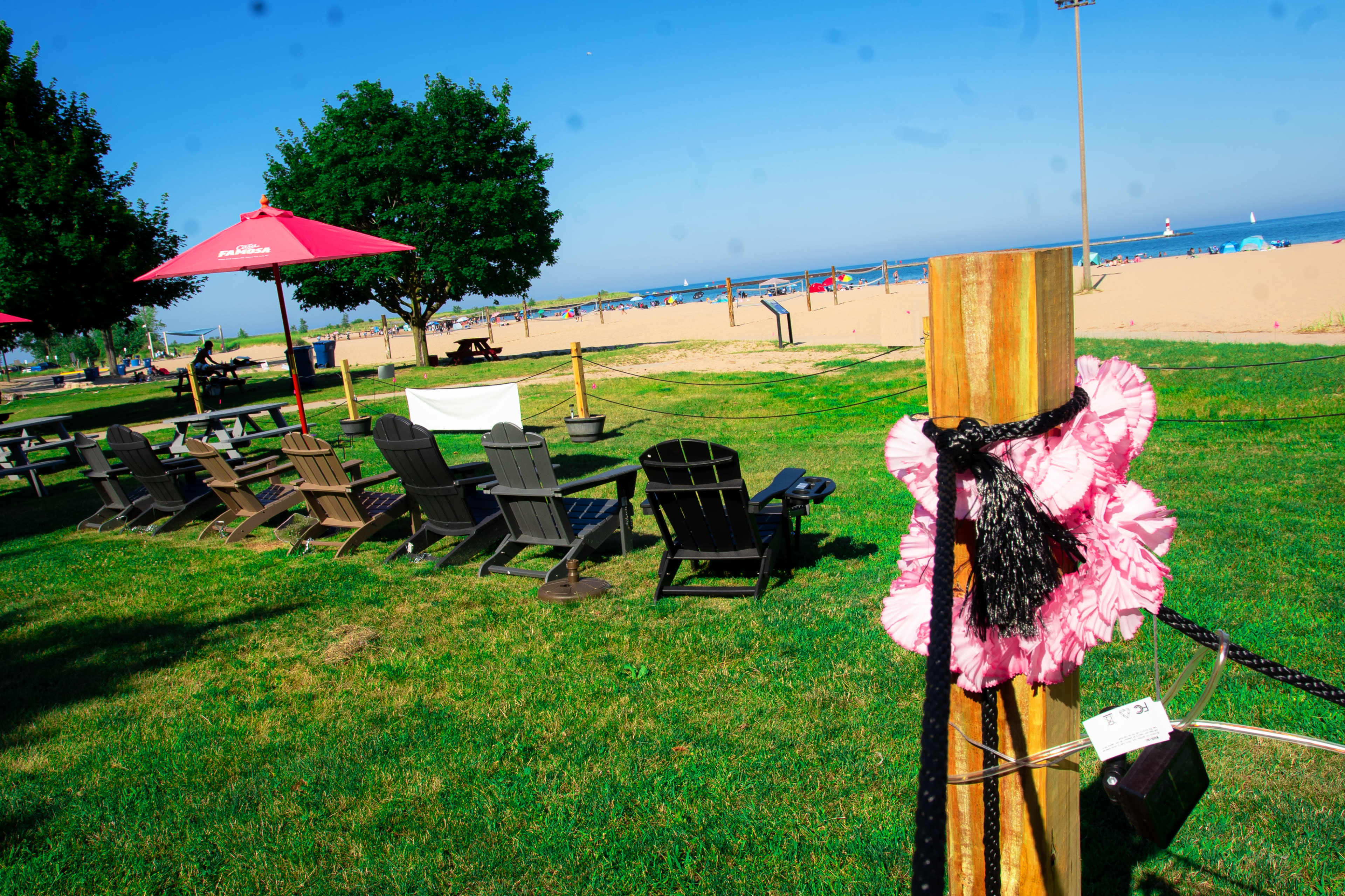 The image shows a grassy area with wooden picnic tables and beach umbrellas, leading to a sandy beach and the ocean in the background.
