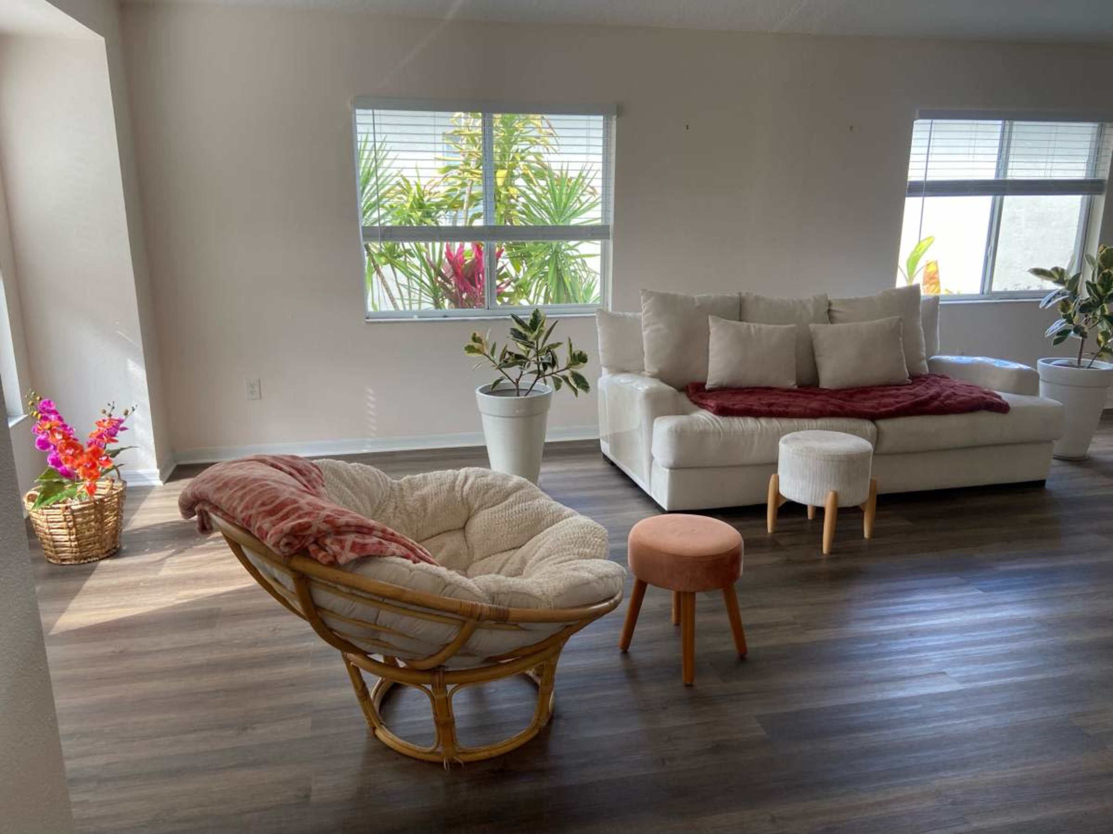 The image shows a bright living room featuring a white sofa with cushions, a round orange stool, a large cozy chair, and potted plants beside windows.