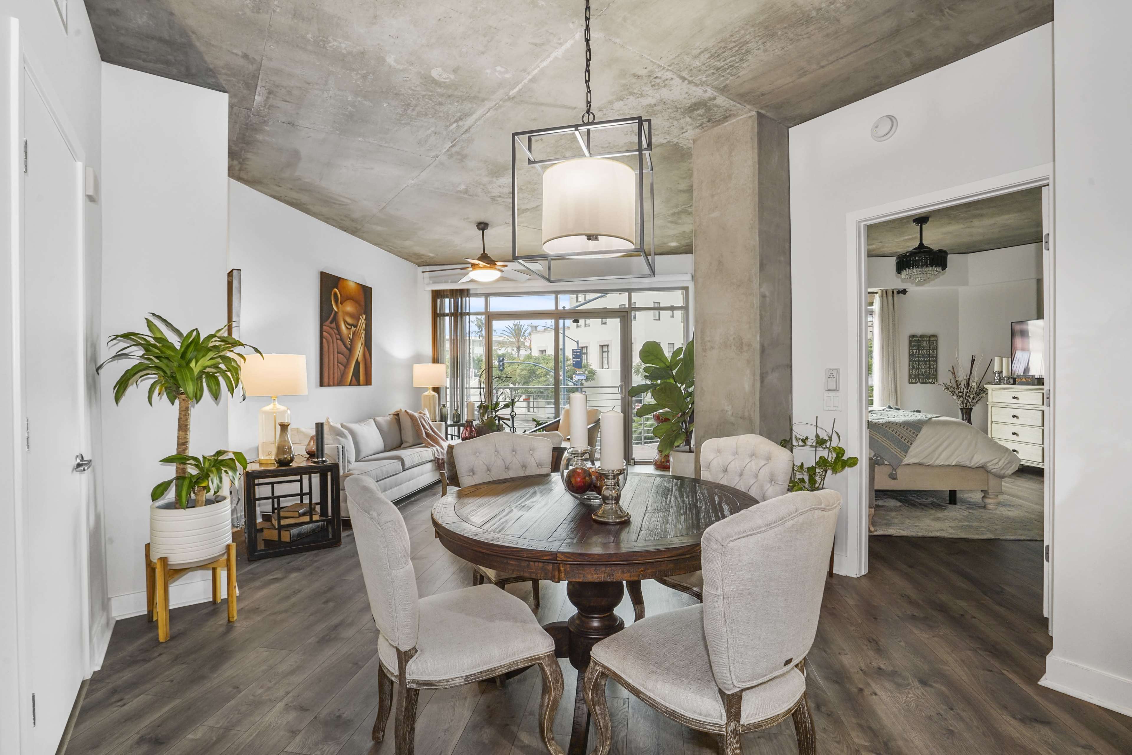 A modern living space features a round dining table surrounded by upholstered chairs, with a seating area visible in the background and large windows letting in natural light.