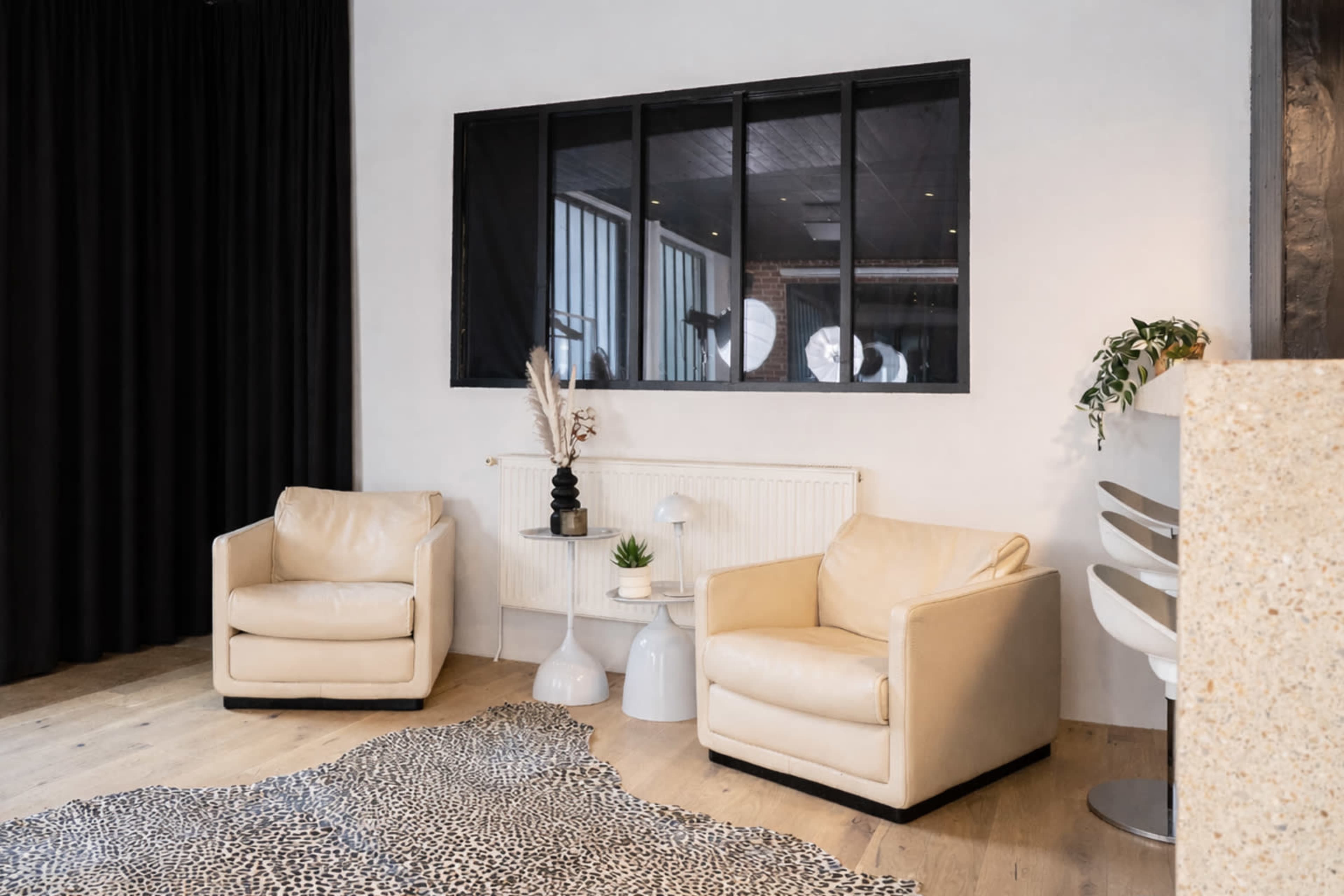 The image shows a modern interior space with two cream-colored armchairs, a round table, and a small plant, all set against a backdrop of dark curtains and a large window.