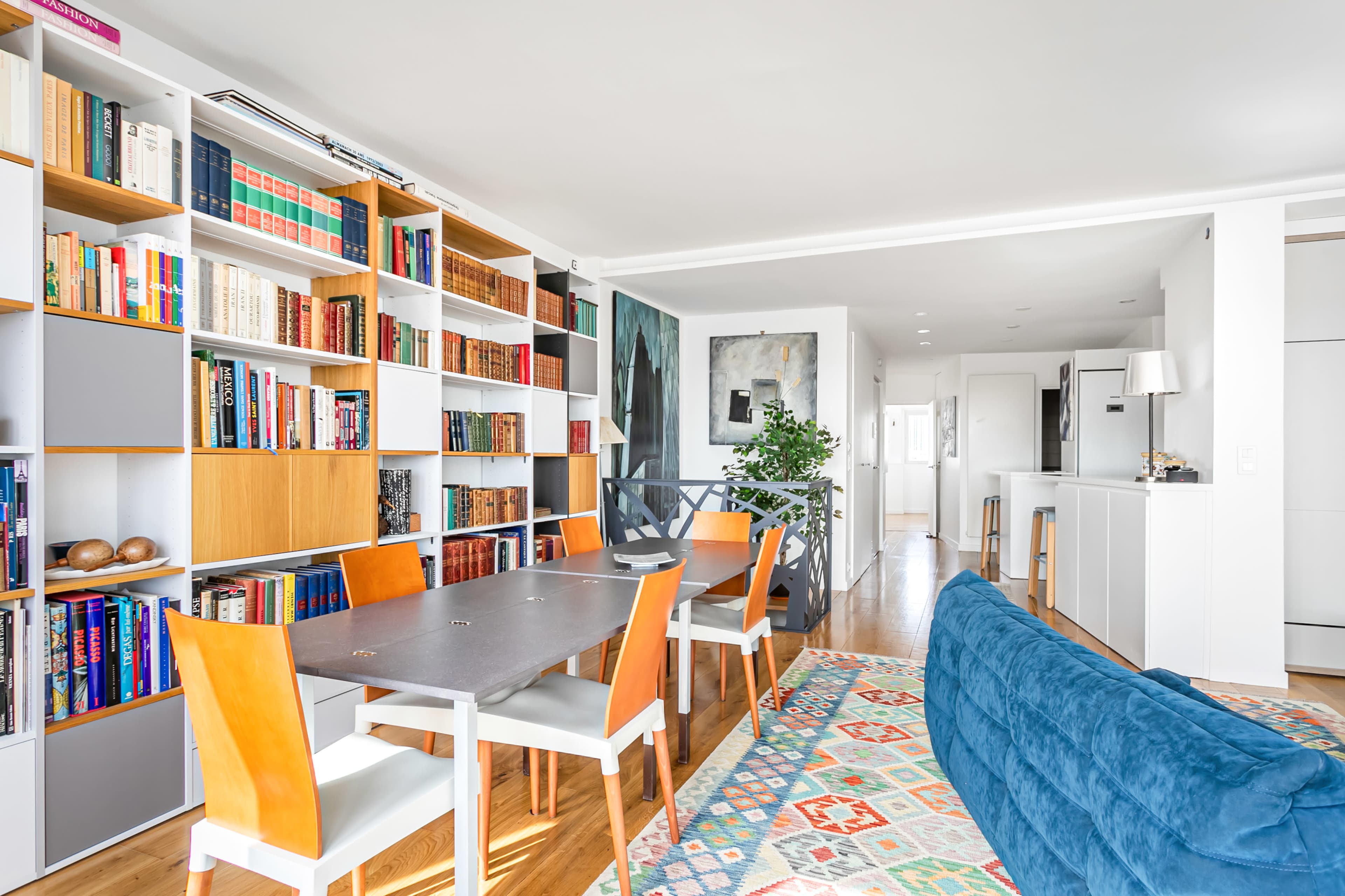 A modern living space features a dining area with a long table and orange chairs, flanked by a bookshelf filled with books and a blue couch.