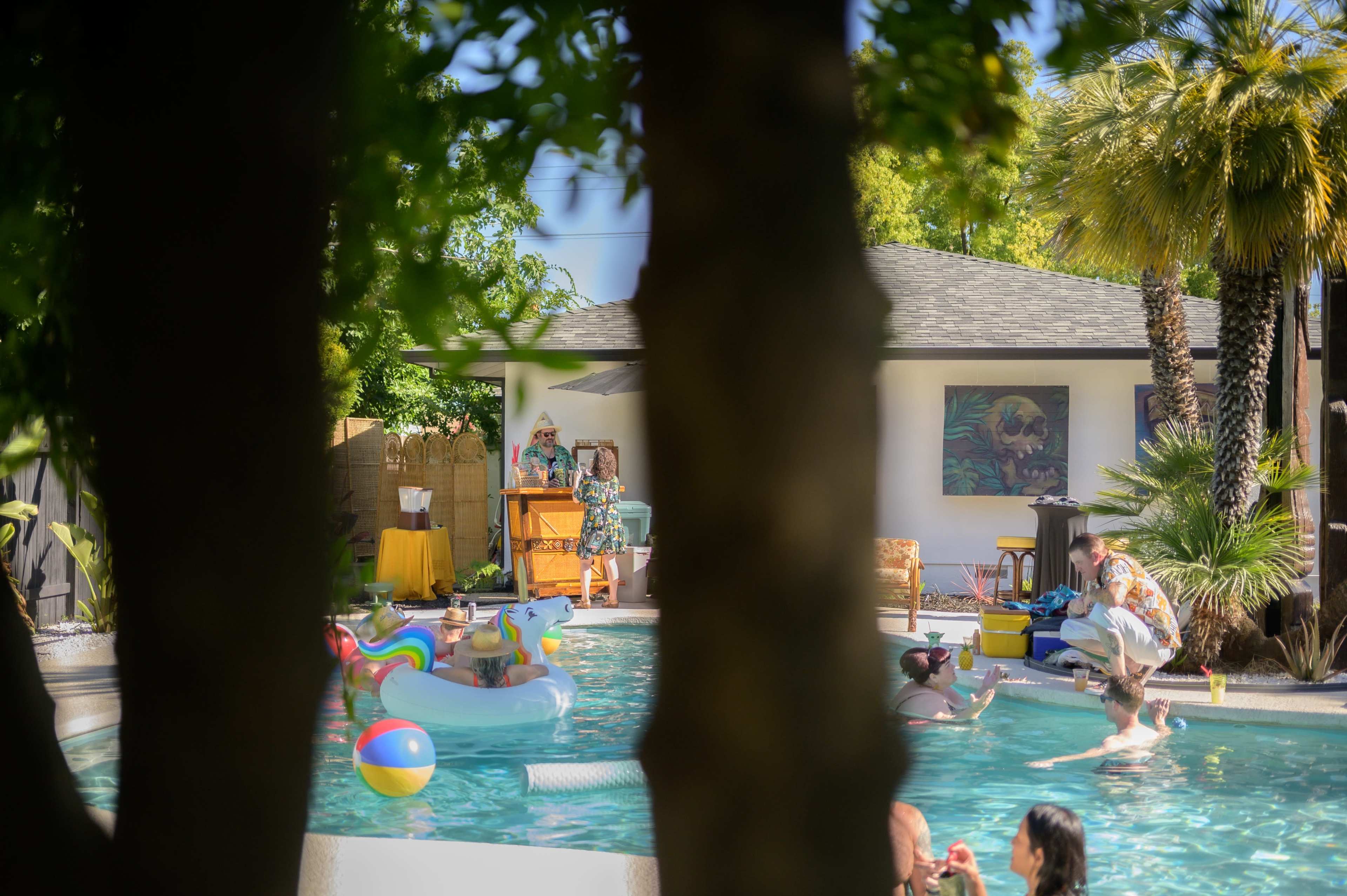 A group of people are enjoying a sunny day at a swimming pool, with colorful floats and lounge chairs around.