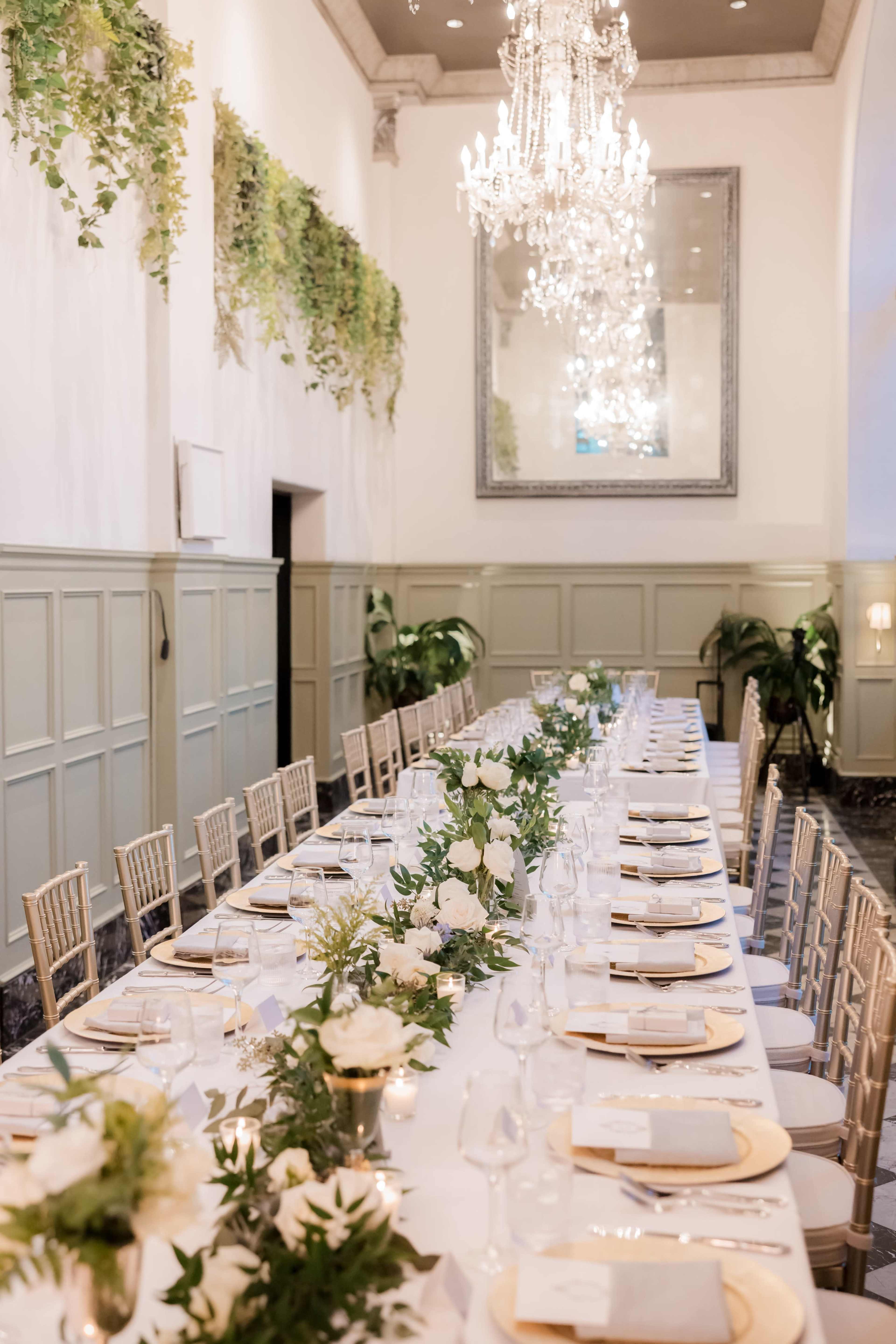 A long banquet table is set with white plates, glassware, and floral arrangements in a elegantly decorated room featuring chandeliers and greenery along the walls.