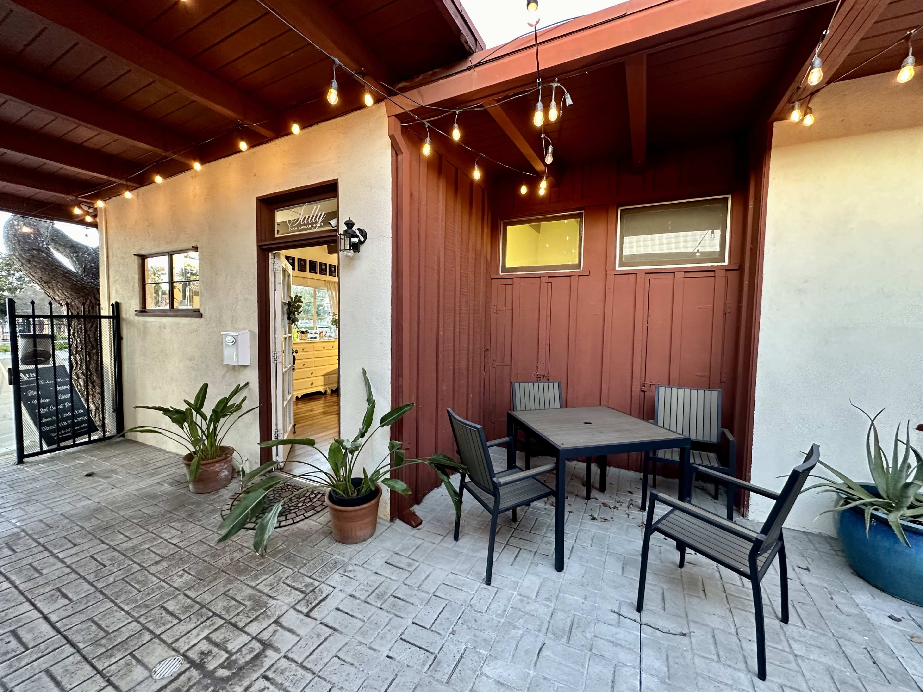 The image shows a small outdoor seating area with a table and chairs, surrounded by potted plants and string lights, adjacent to a building entrance.