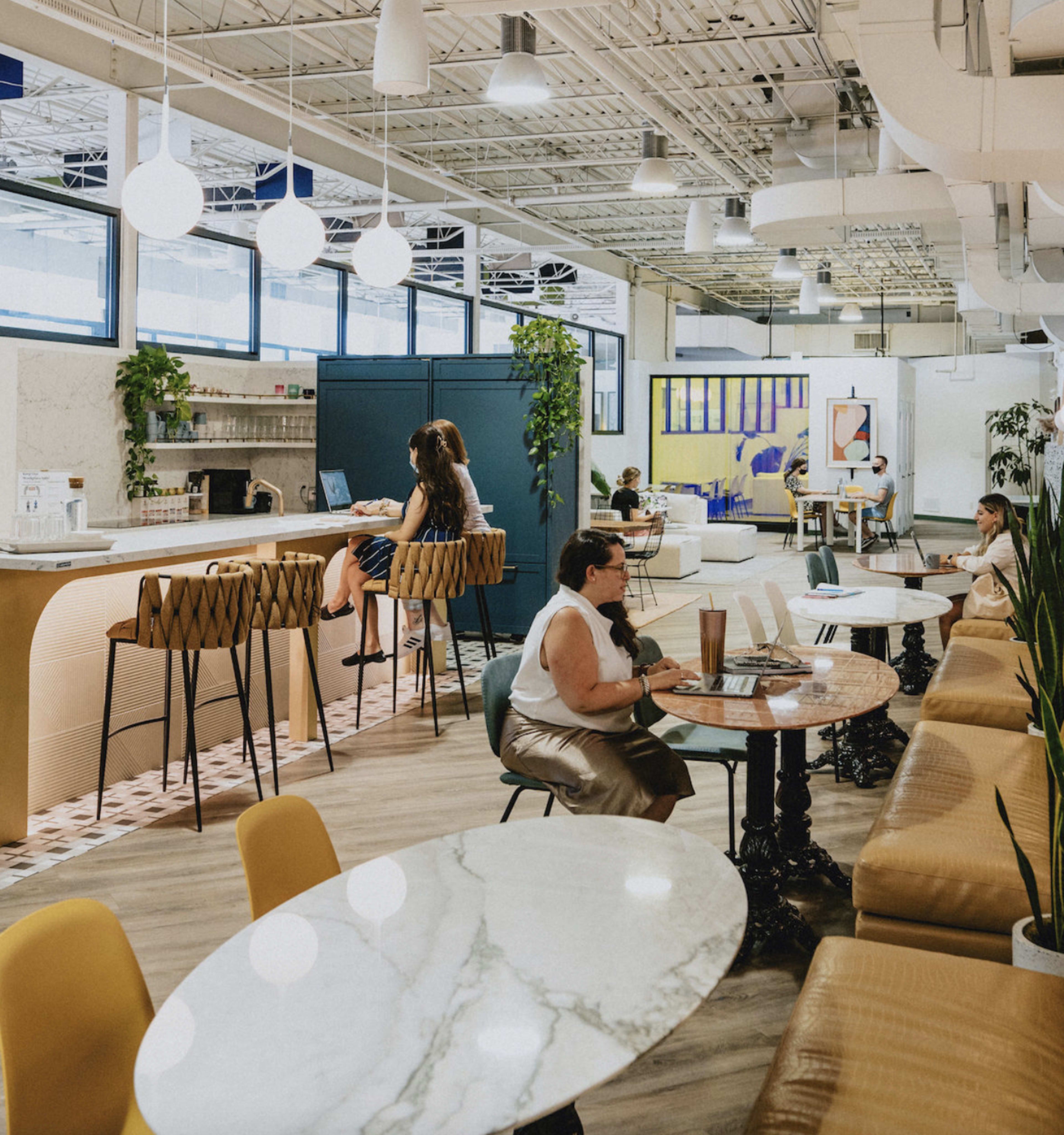 A modern co-working space features a variety of seating arrangements, including high stools at a bar and tables with chairs, where individuals are engaged in work.