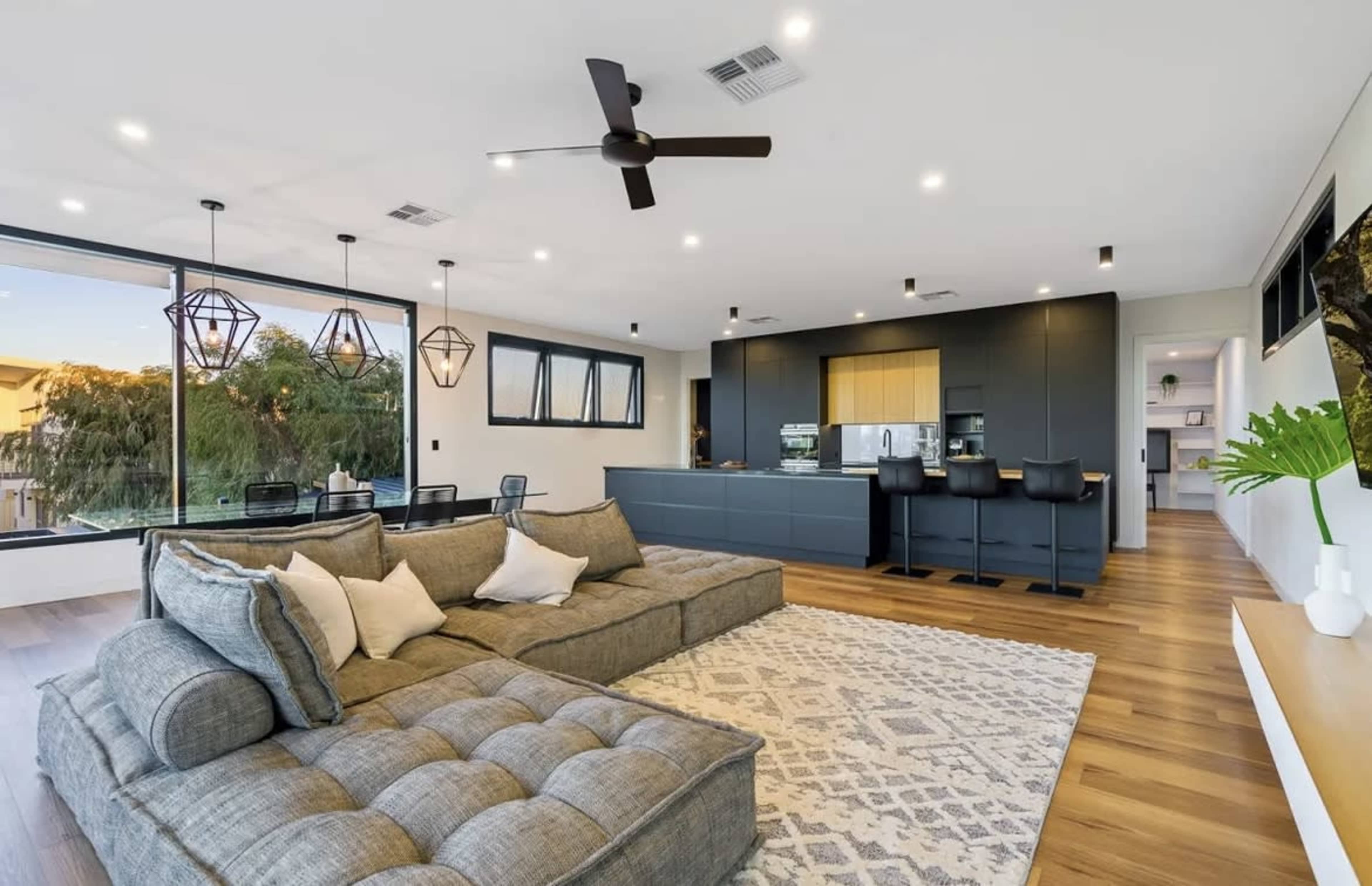 The image shows a modern living room featuring a large sectional sofa, pendant lighting, and a sleek kitchen in the background.