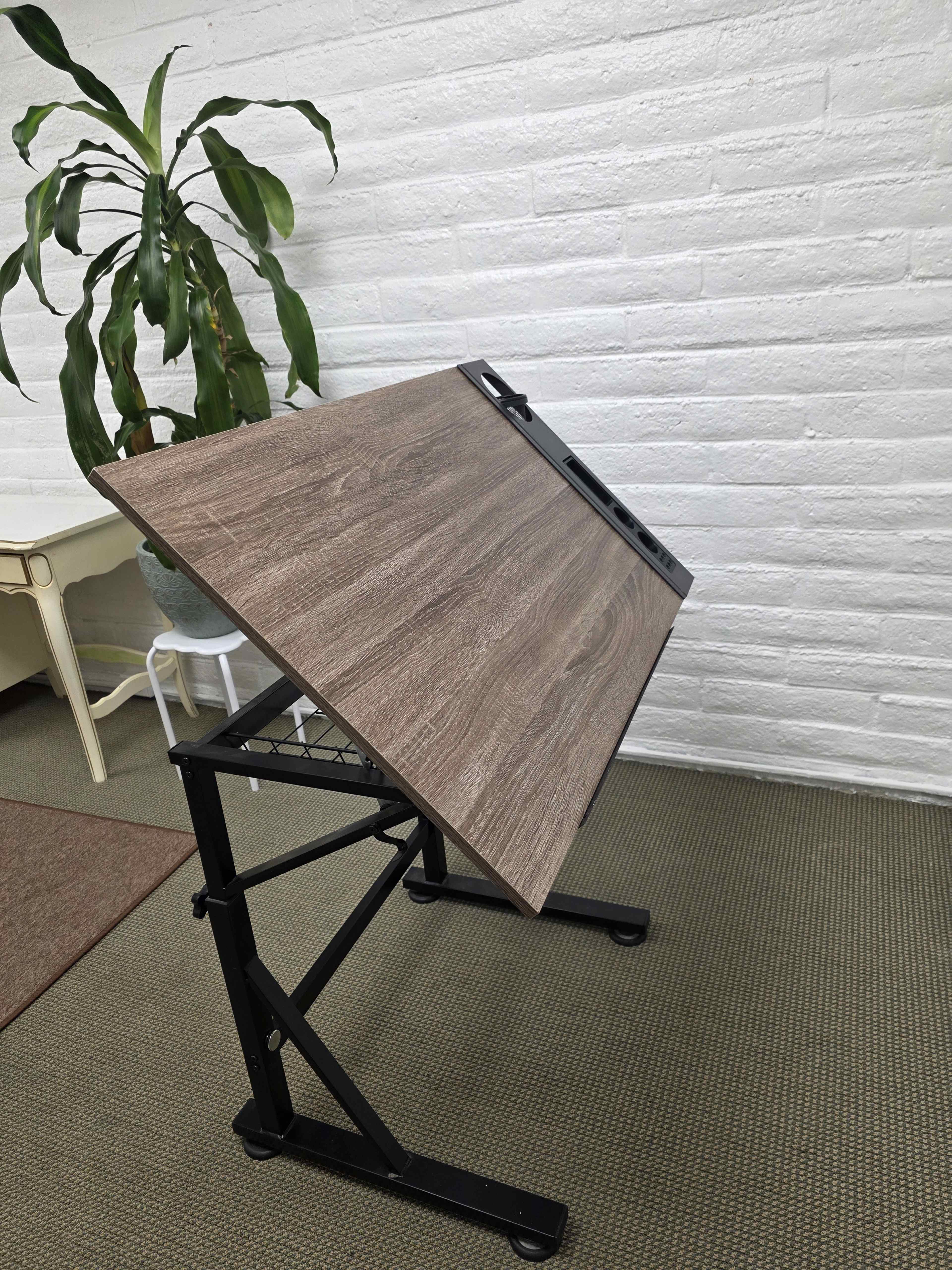 The image shows an adjustable drafting table made of wood with a dark finish, positioned in a room with white brick walls and a large green plant in a pot nearby.